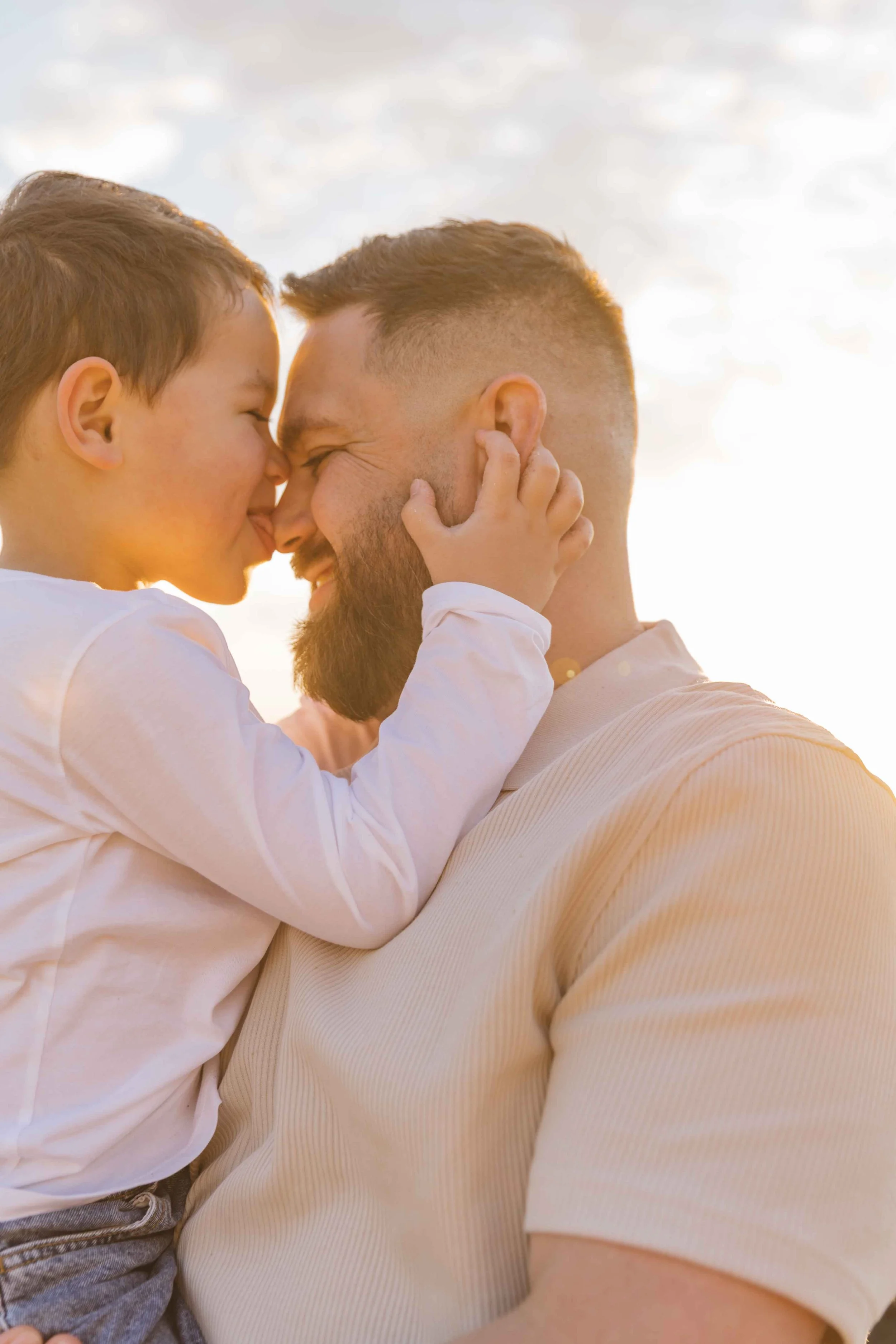 A young boy and an adult man, possibly father and son, face each other nose to nose with their eyes closed. The boy's hands gently touch the man's face, and they are outdoors with a cloudy sky in the background, sharing a joyful moment.