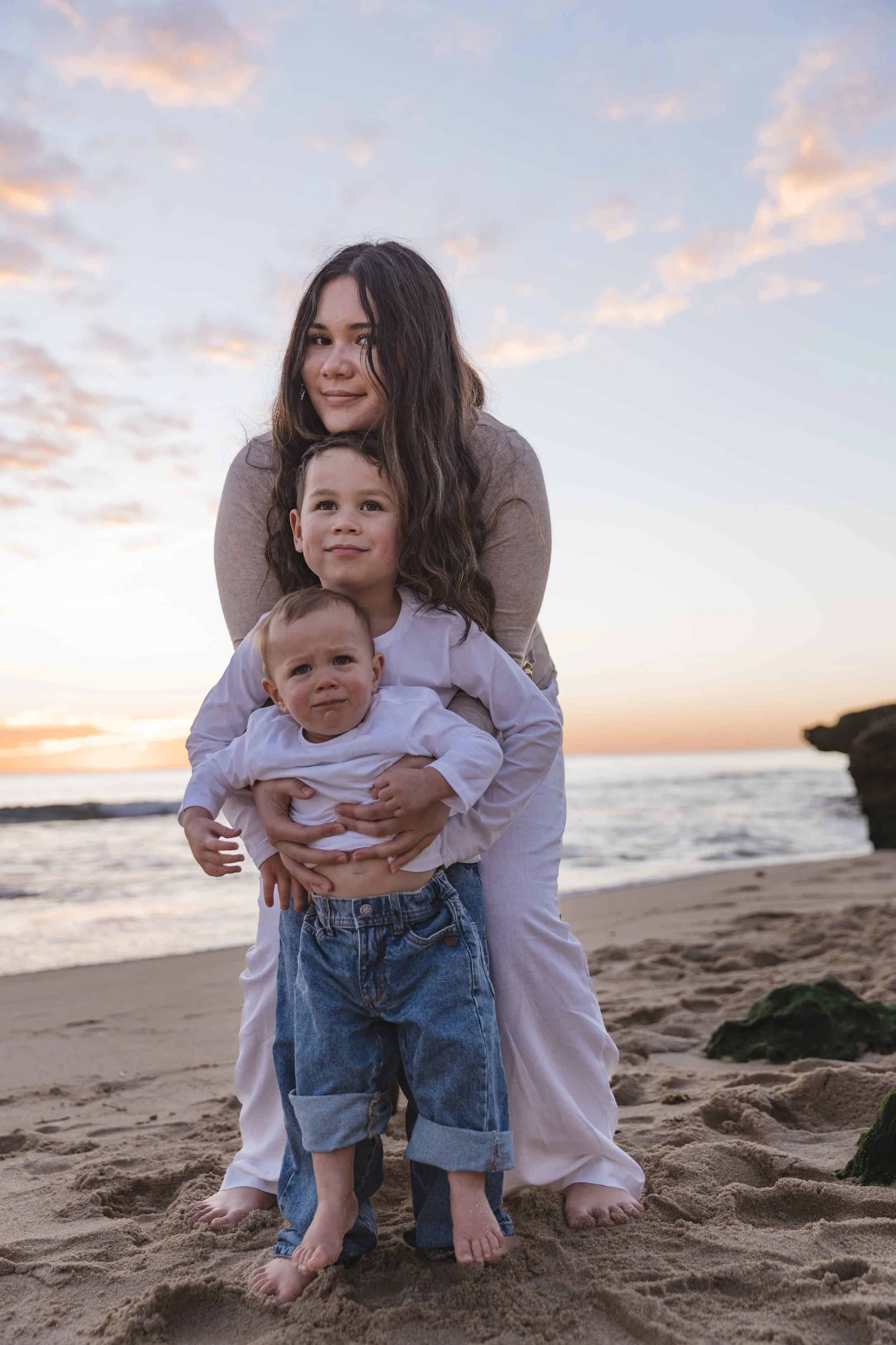 Three siblings, standing on a beach at sunset, with ocean and rocks in the background.