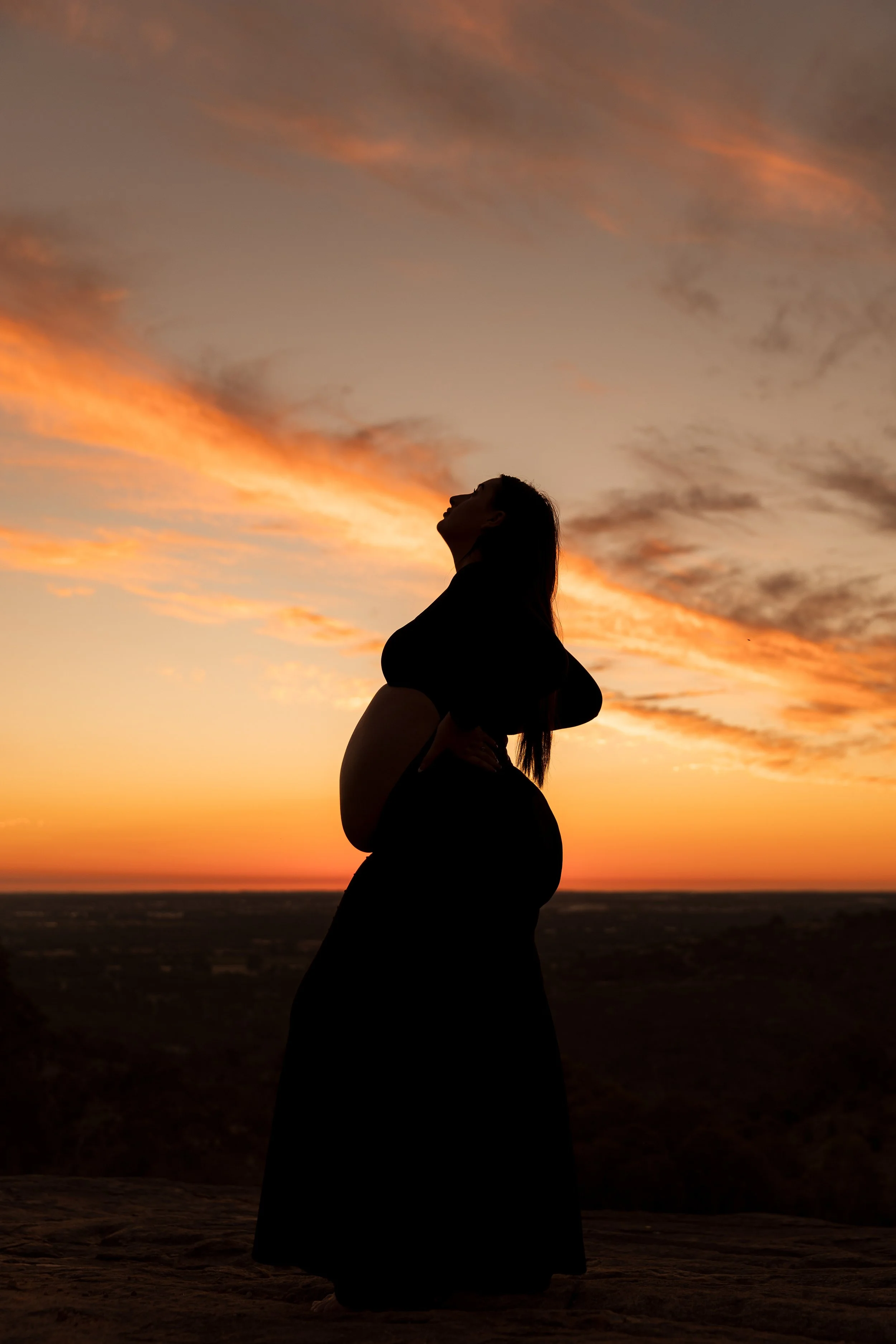 Silhouette of a pregnant woman outdoors during sunset, standing with her hands on her stomach, with a colorful sky and clouds in the background.