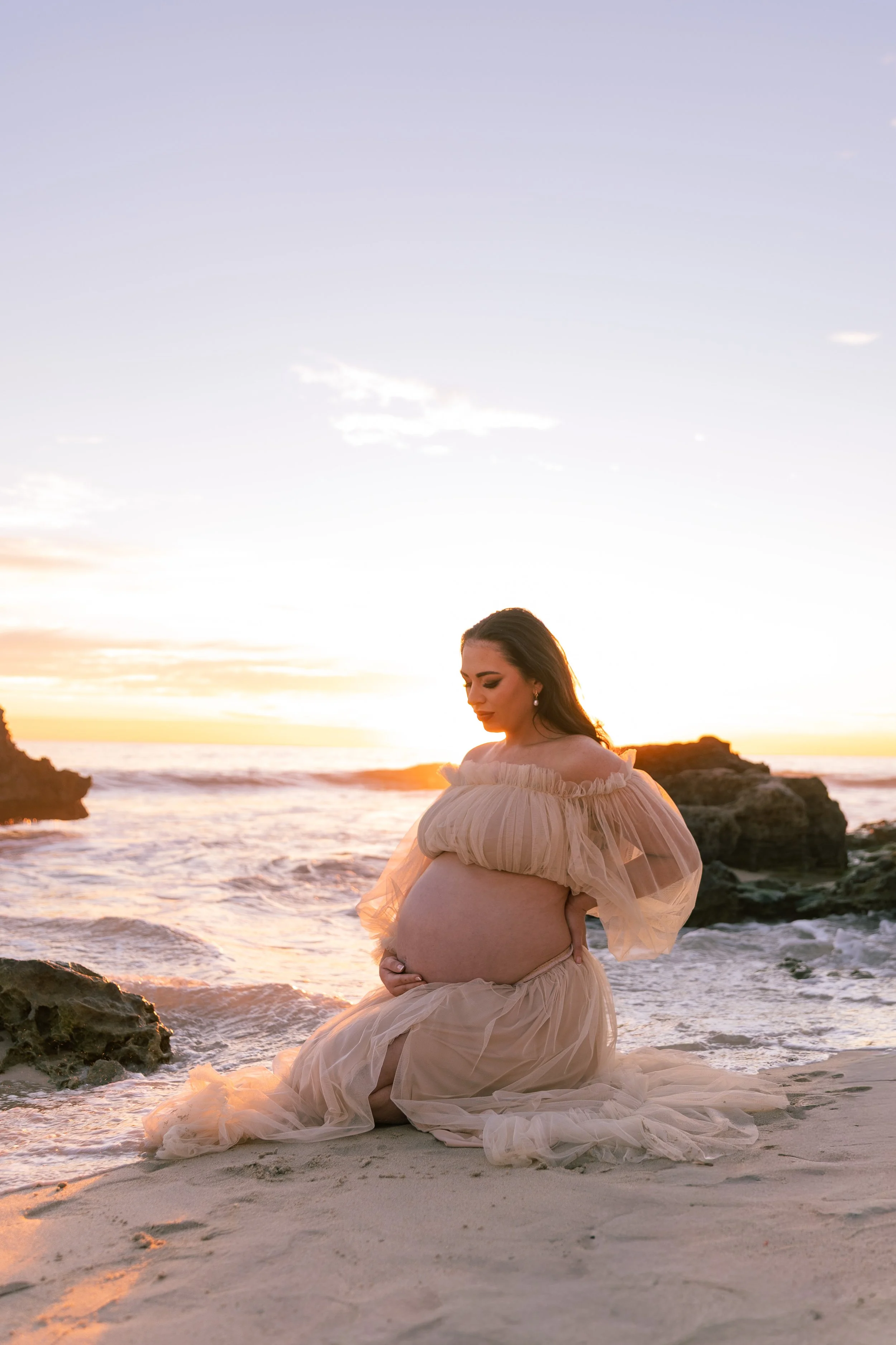 A pregnant woman in a flowing beige dress kneels on a beach at sunset, holding her belly with her eyes closed, near rocks and the ocean.