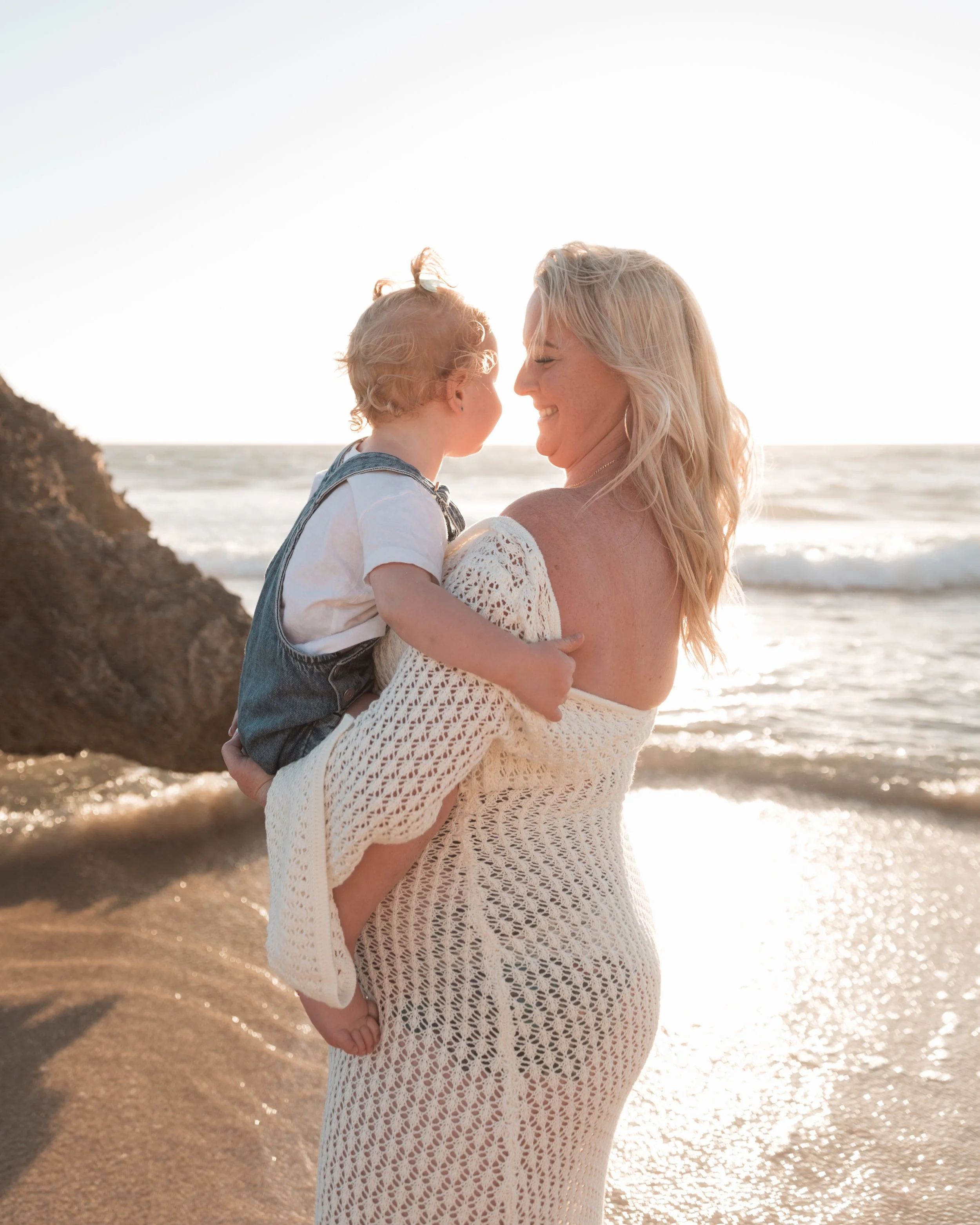 A woman with long blonde hair holding a young child with blonde curly hair at the beach during sunset, near the shoreline with waves and a rocky outcrop in the background.