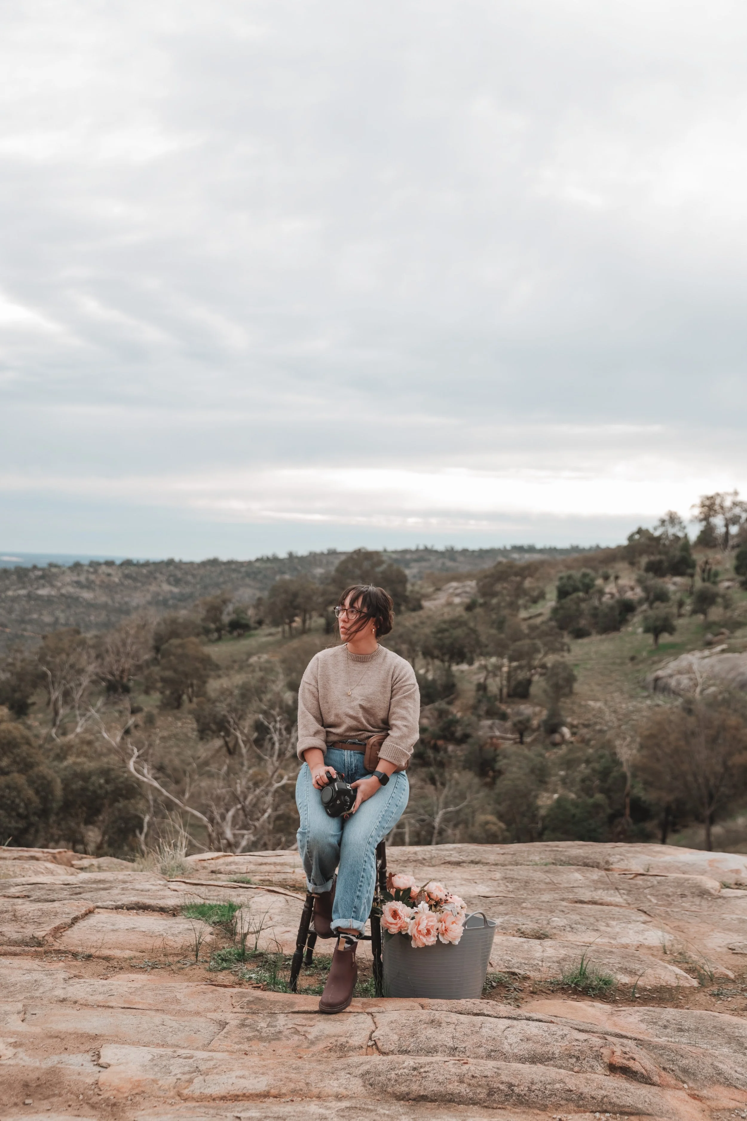A woman sitting outdoors on a large rock, holding a camera, with a bucket of pink flowers beside her, in a scenic landscape with rolling hills, sparse trees, and an overcast sky.