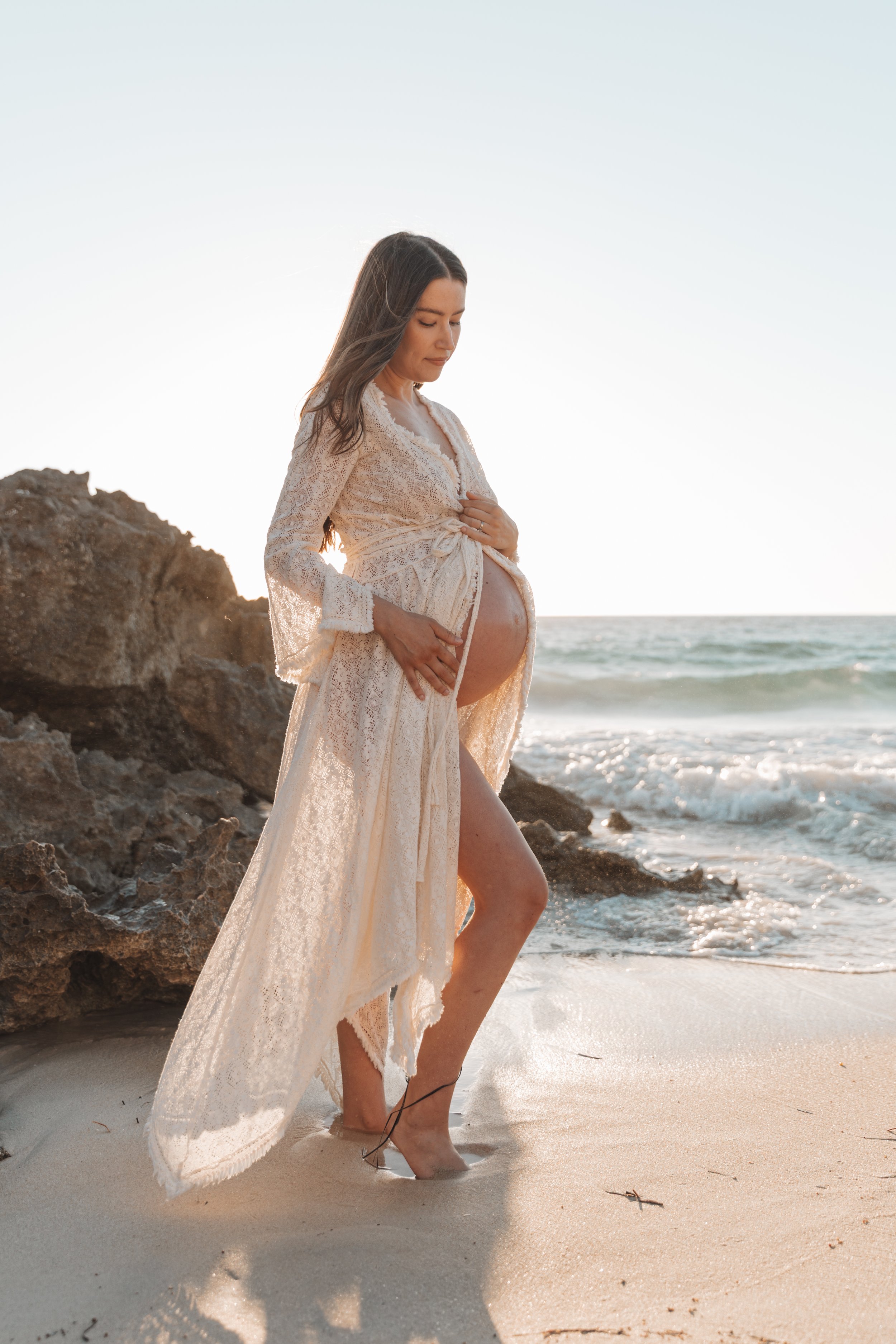 Pregnant woman standing barefoot on the beach near rocks, wearing a delicate white lace dress and holding her belly, with the ocean and sky in the background.