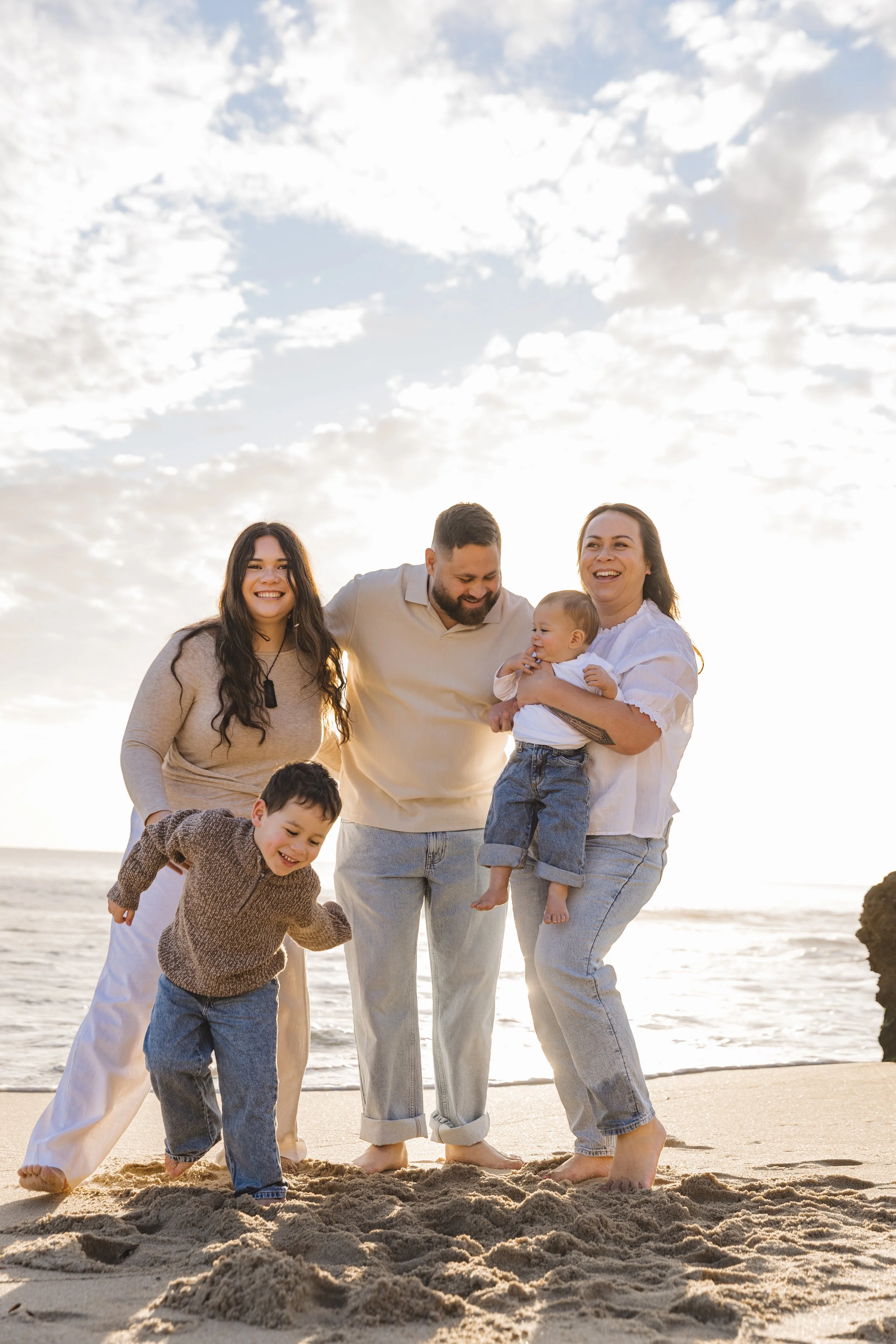 A family of six enjoying time on the beach, including two children, standing in the sand and smiling, with a cloudy sky overhead.