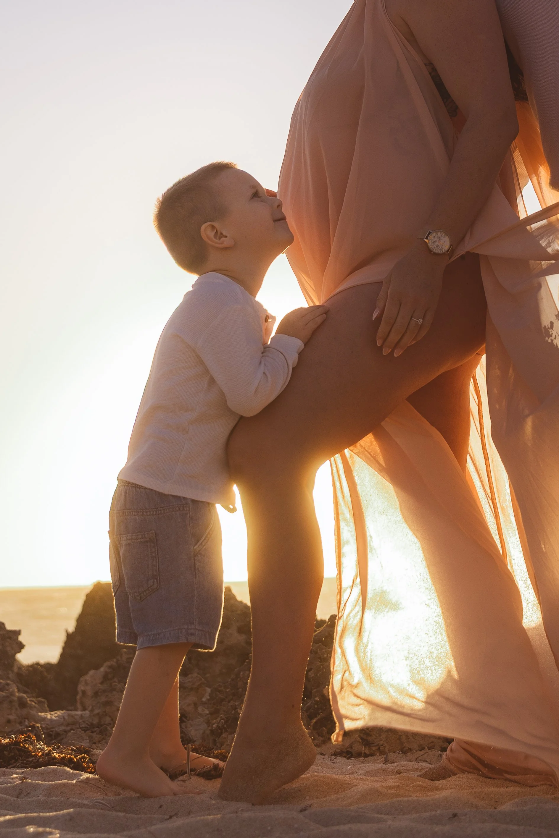 A young boy and a woman, likely his mother, standing on a sandy beach at sunset. The boy is smiling and looking up at the woman, who is touching her pregnant belly. The scene is warm and intimate, with the ocean and rocks visible in the background.