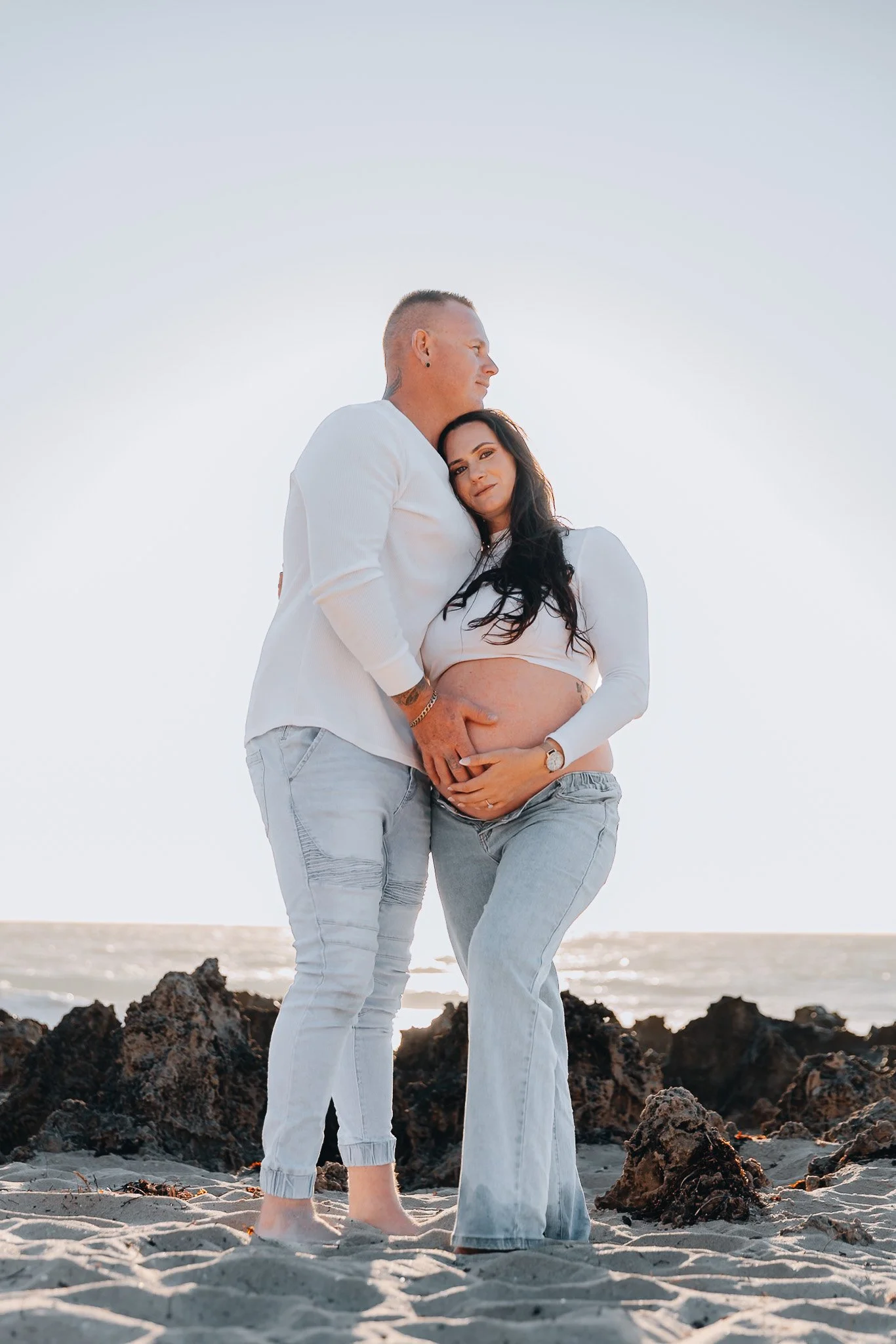 A couple standing on a rocky beach near the ocean, with the woman showcasing a pregnant belly, and both wearing white clothing, during sunset or sunrise.