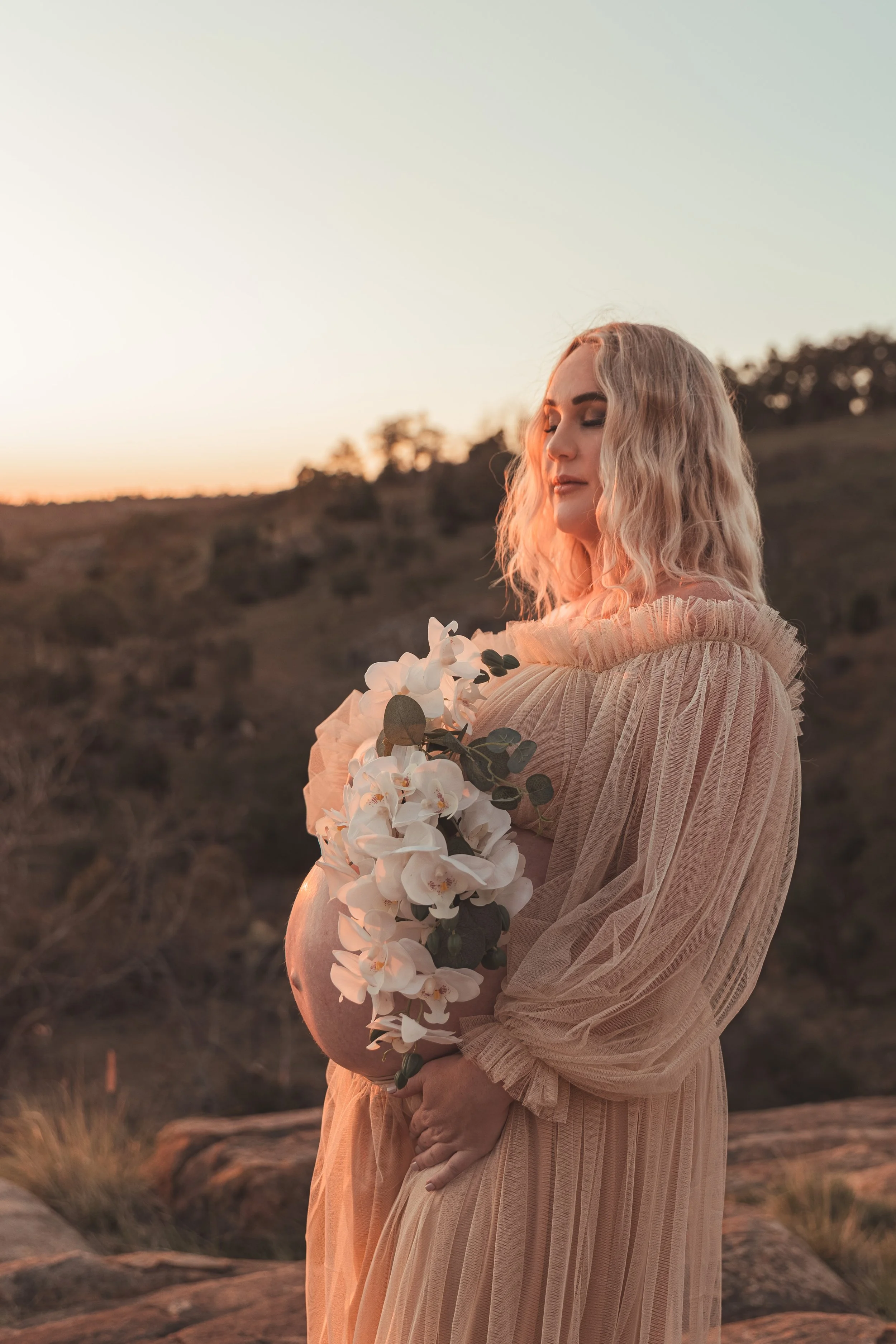 A pregnant woman with wavy blonde hair standing outdoors during sunset, holding a bouquet of white orchids and greenery, wearing a flowing beige dress and with eyes closed.