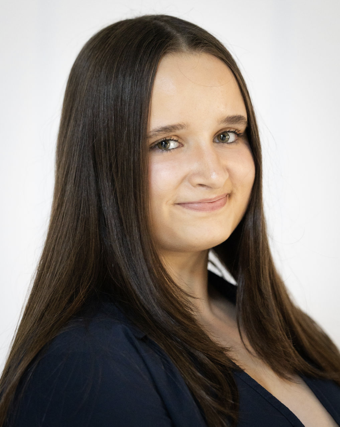 Headshot of a young woman with long brown hair, light skin, and green eyes, smiling slightly, wearing a dark top.