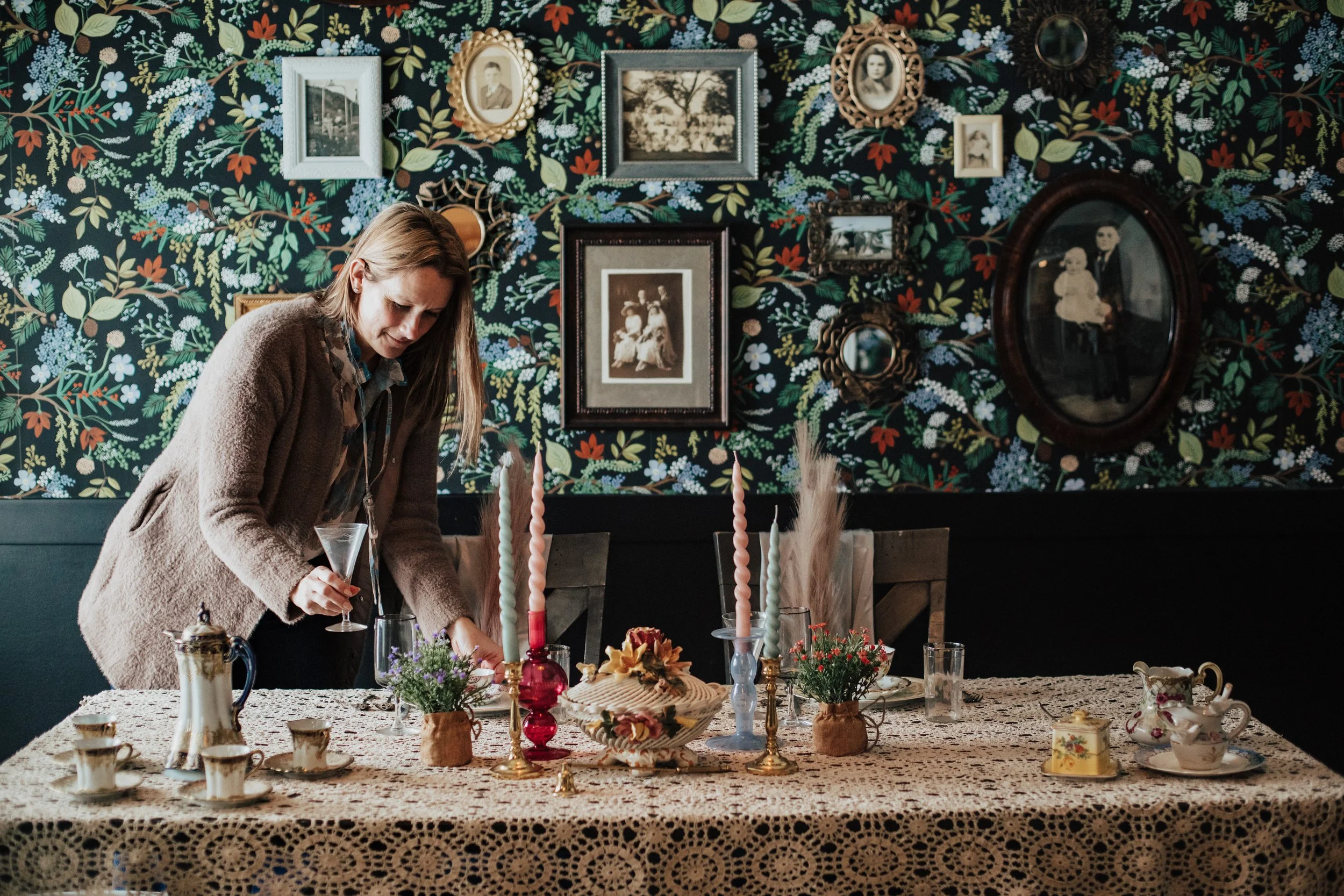A woman dressed in a beige sweater and shirt is standing at a decorated table, preparing drinks. The table has a lace tablecloth, colorful candles, potted flowers, and vintage tea cups. The background has a dark floral wallpaper with framed vintage photographs and mirrors.