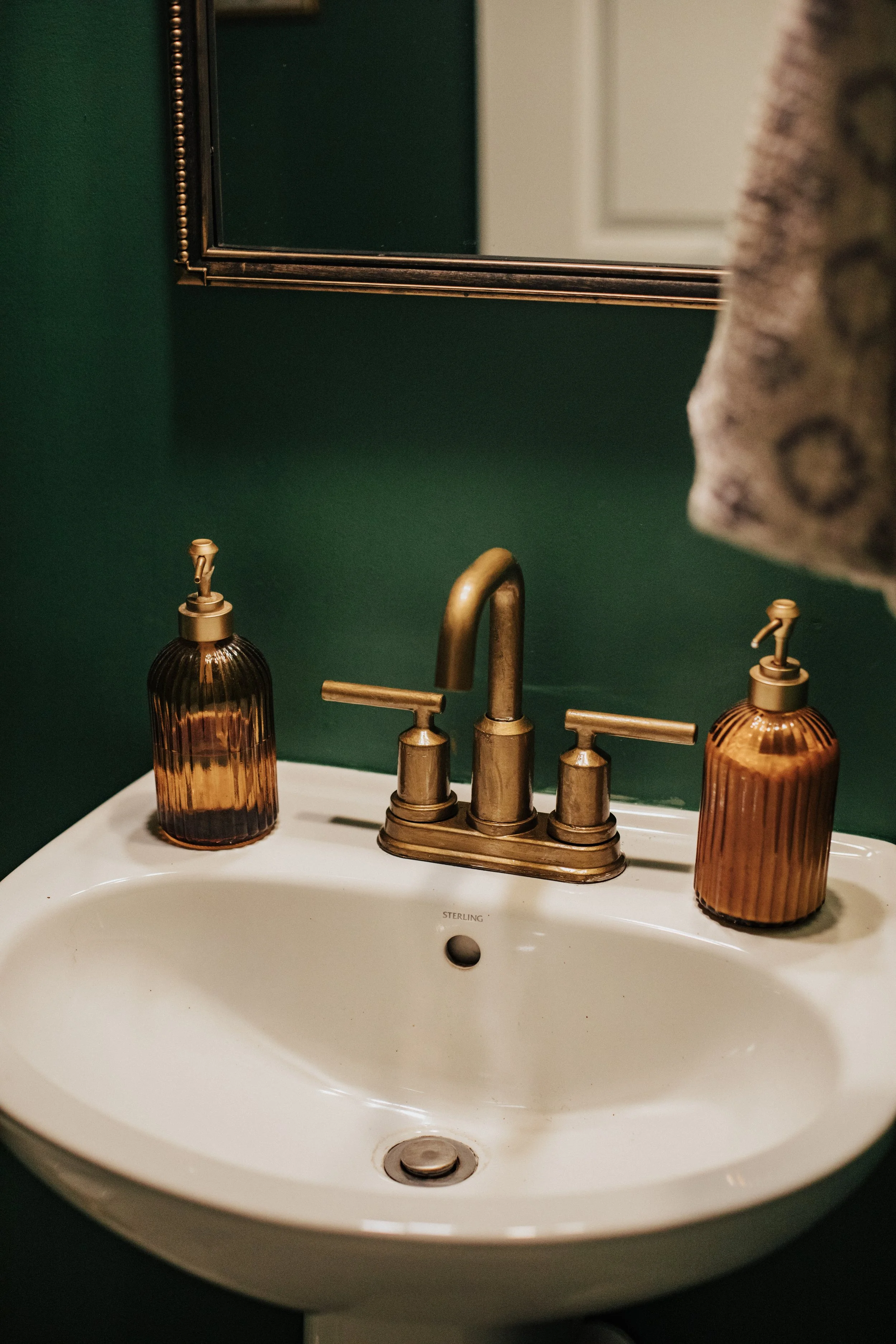 A white bathroom sink with a bronze faucet and two matching soap dispensers, against a dark green wall, with a mirror above.