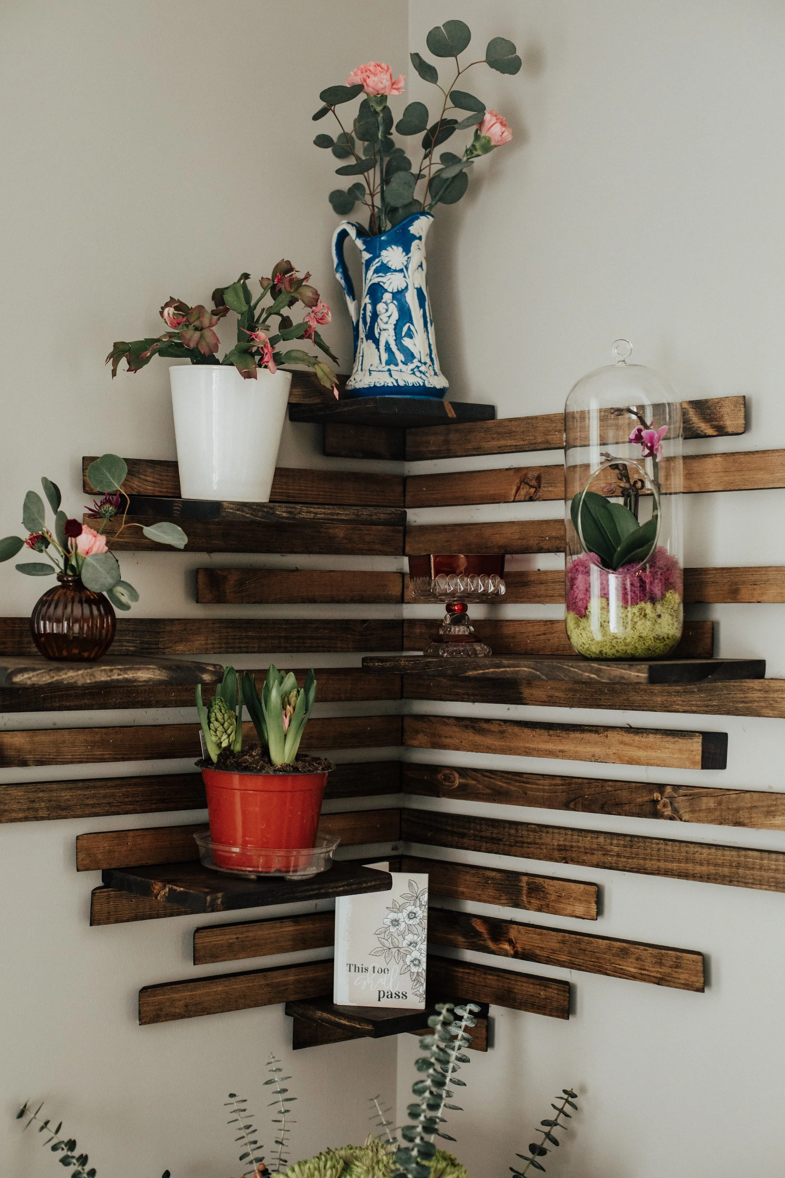 Decorative wall with wooden slats holding various potted plants and vases, including pink flowers and a card with a floral design.