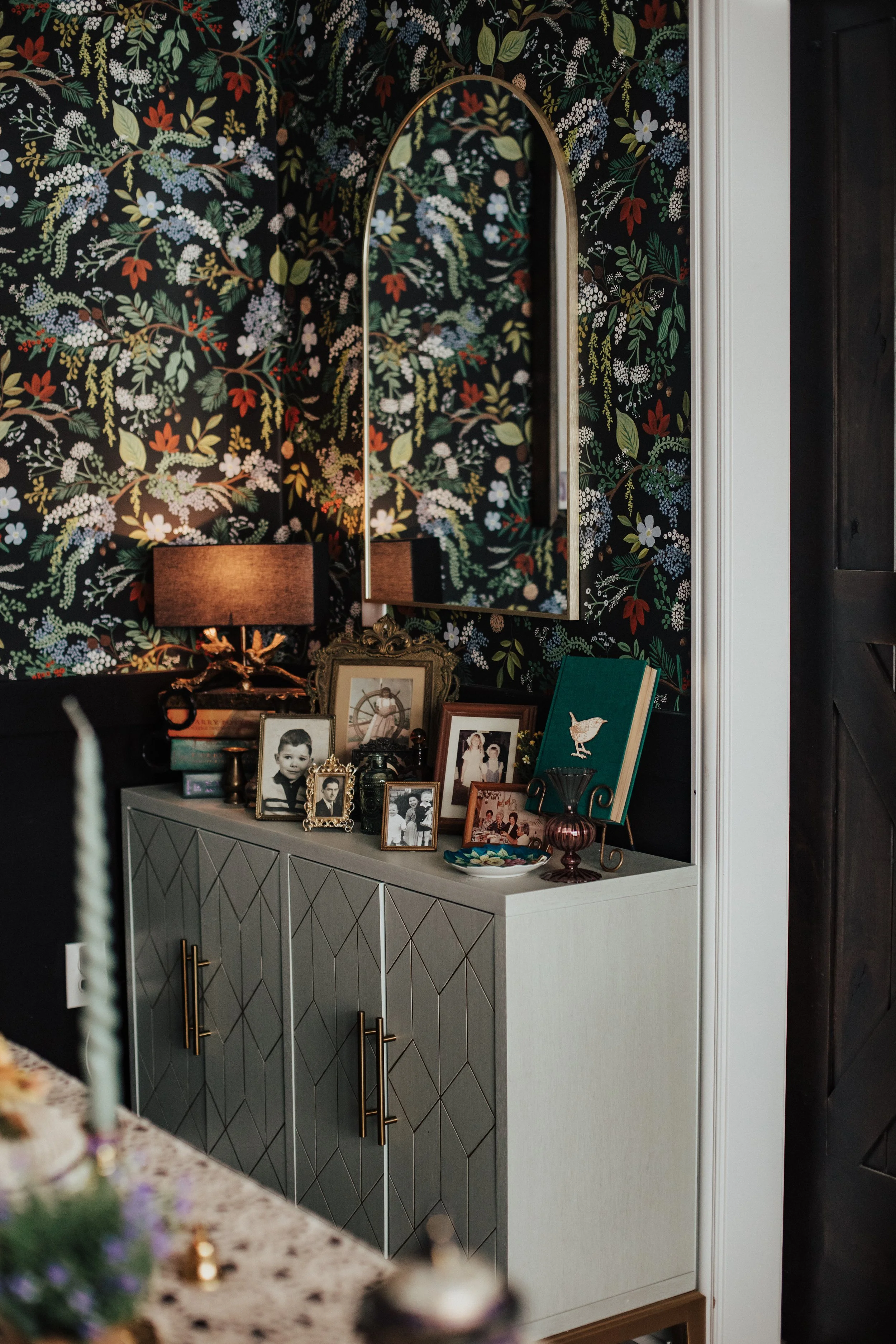 A modern sideboard decorated with framed black-and-white photographs, a decorative book with a bird on the cover, a small green plant, a purple glass vase, and a bowl with candies. Above it, a large, arched mirror reflects the floral wallpaper behind. The wallpaper features a dark background with colorful floral patterns, and there are two table lamps with dark shades on either side of the mirror.