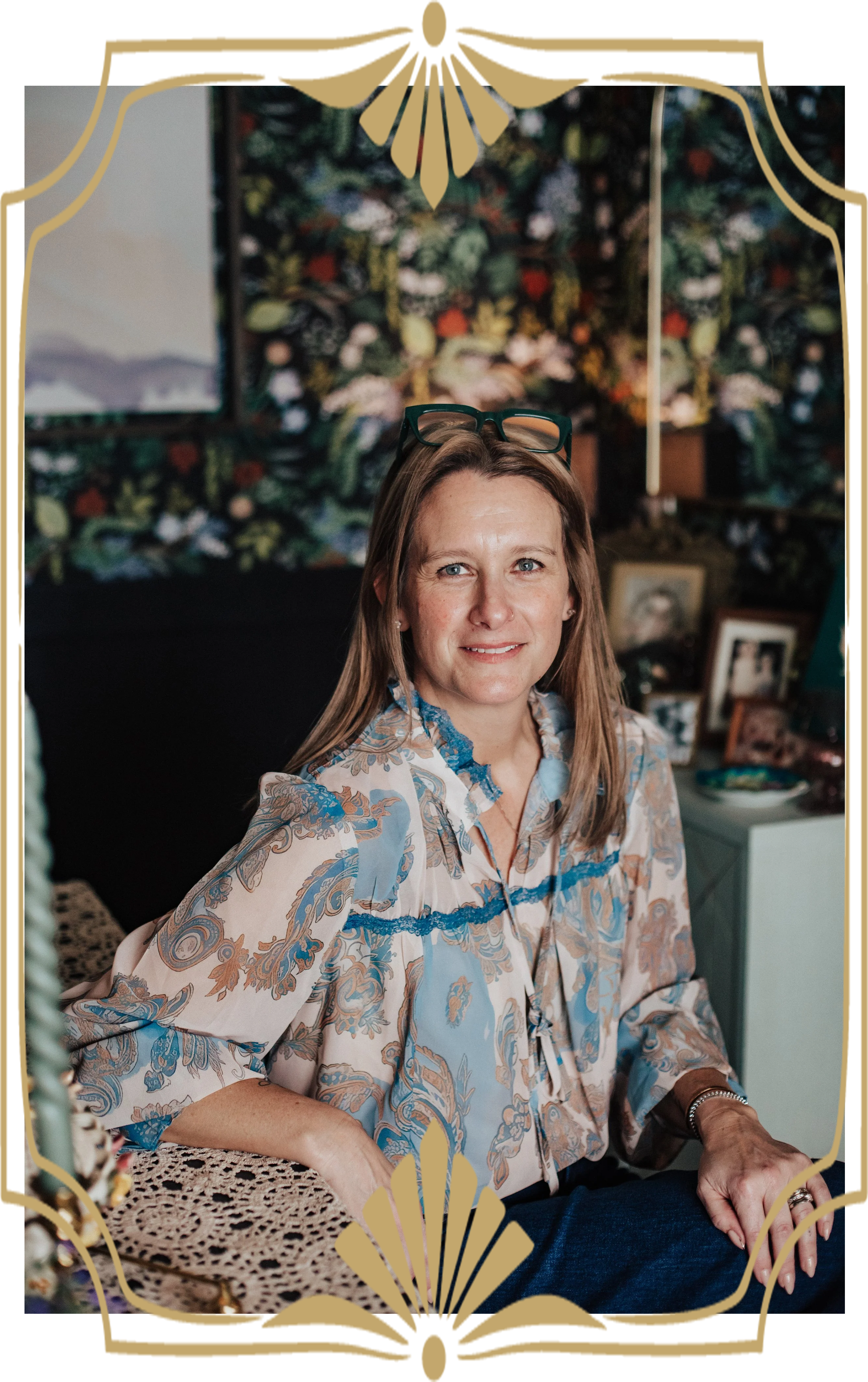 A woman sitting at a table with a lace tablecloth, smiling, wearing a patterned blouse with blue and beige designs, glasses resting on her head, in a room with floral wallpaper and framed photos.