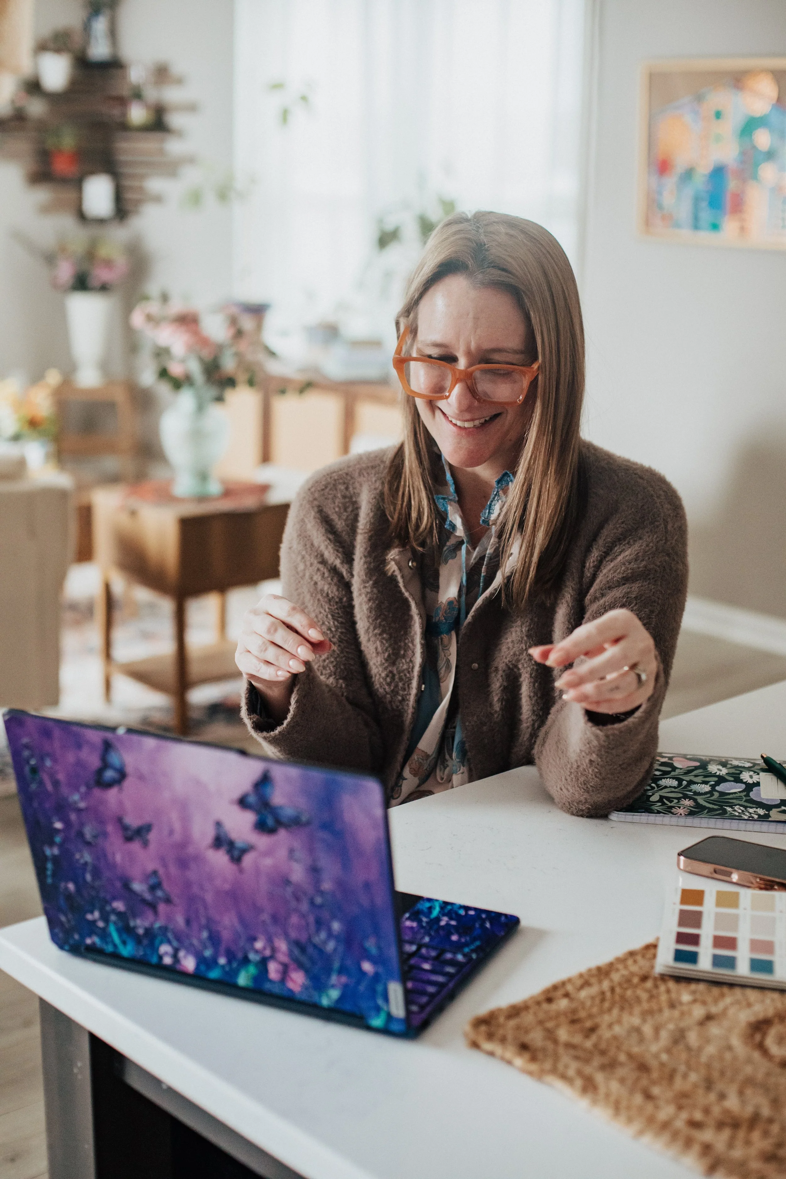 A woman with long brown hair, glasses, and a brown sweater smiling and pointing while sitting at a desk with a purple butterfly-themed laptop in a decorated and bright living room.