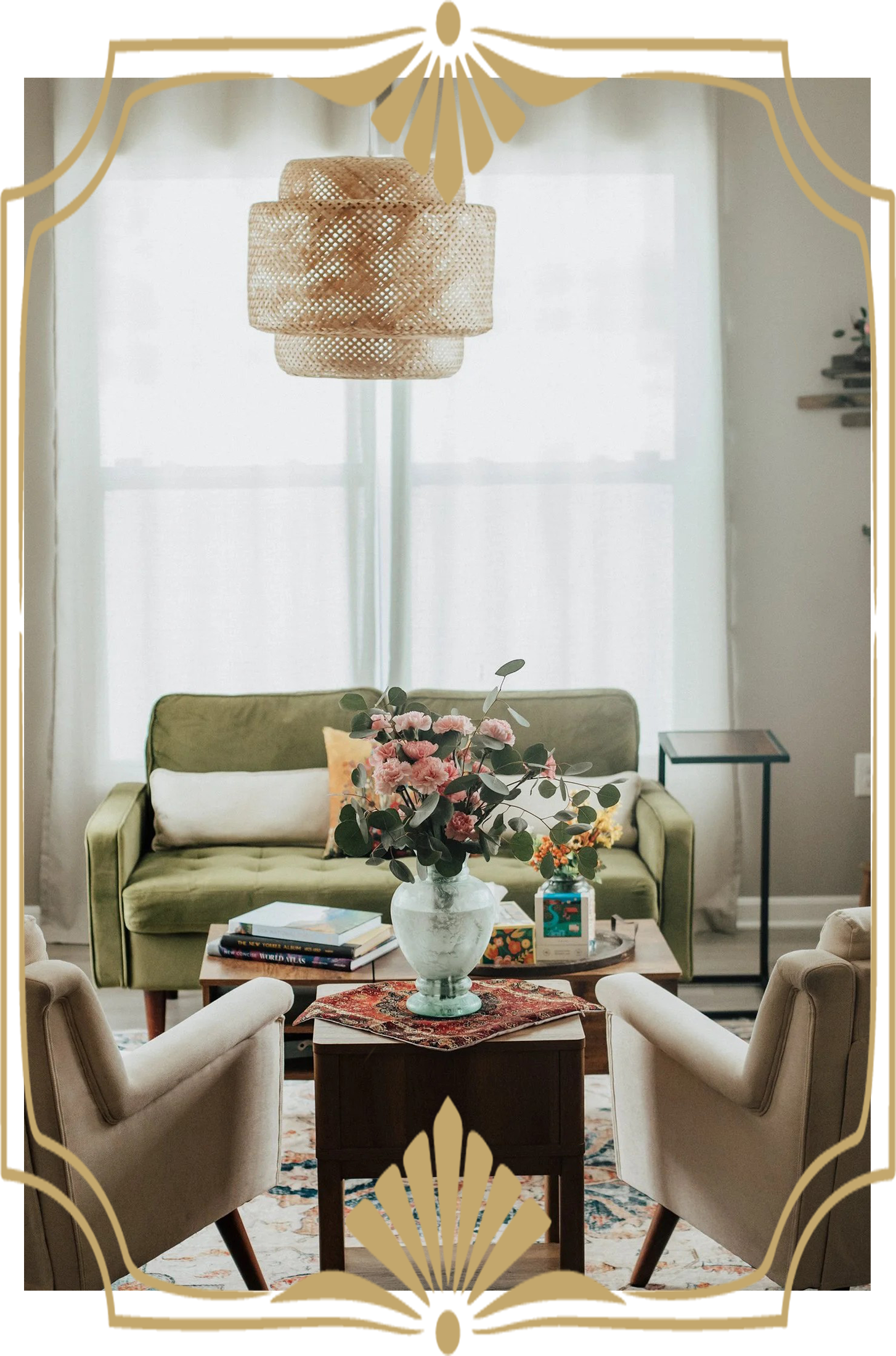 Living room with a green sofa, upholstered chairs around a coffee table with a floral centerpiece, books, and decorative items, curtains and a window in the background, and a woven pendant light hanging from the ceiling.
