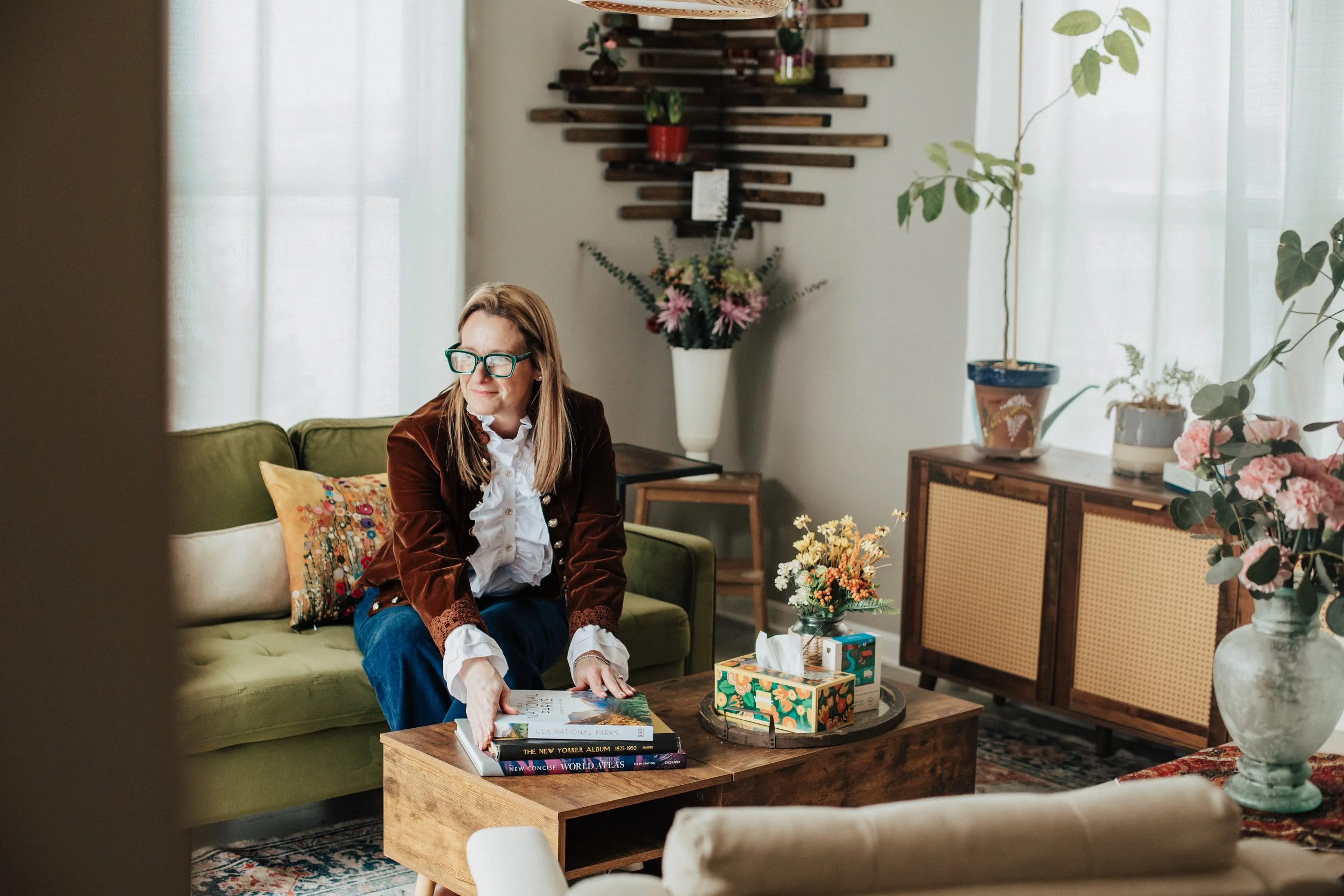 A woman sitting on a green sofa in a living room, wearing glasses, a brown blazer, and white blouse, looking to her right, with a coffee table in front of her holding books, tissues, and a decorative tissue box. The background features potted plants, a wooden wall art, and sheer curtains.