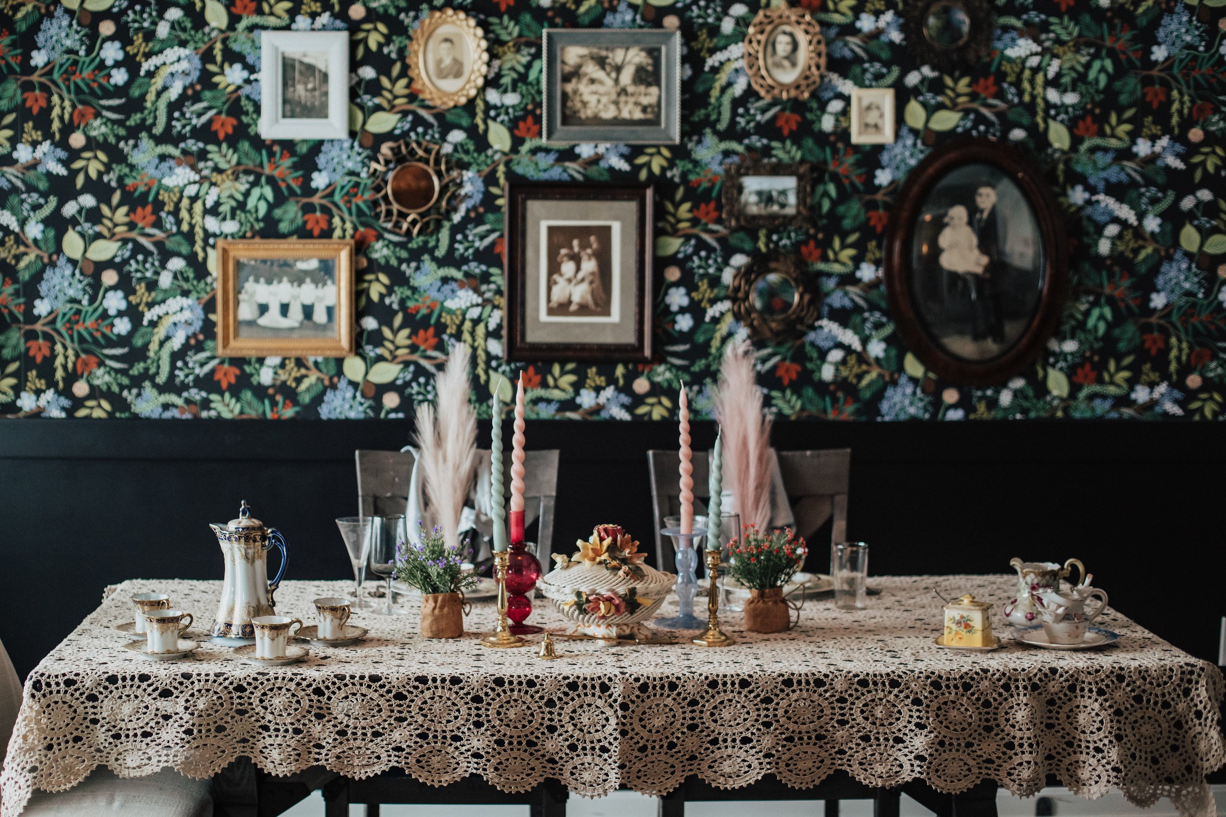 A dining table decorated with a lace tablecloth, candles, teapot set, and floral arrangements in a room with floral wallpaper and vintage framed photographs on the wall.