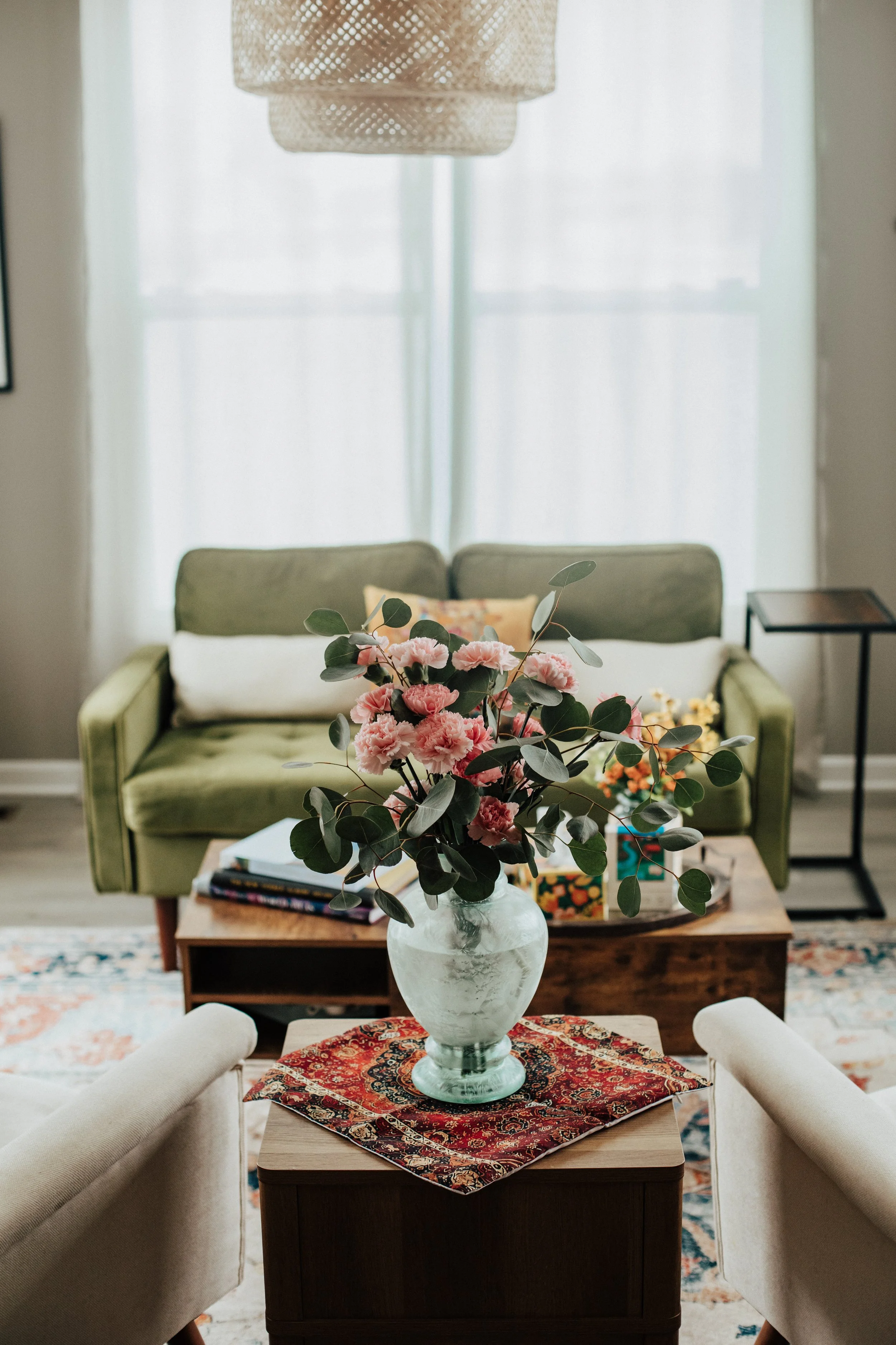 Living room with green sofa, beige armchair, coffee table with pink flowers in a glass vase, patterned rug, and large window with sheer curtains.