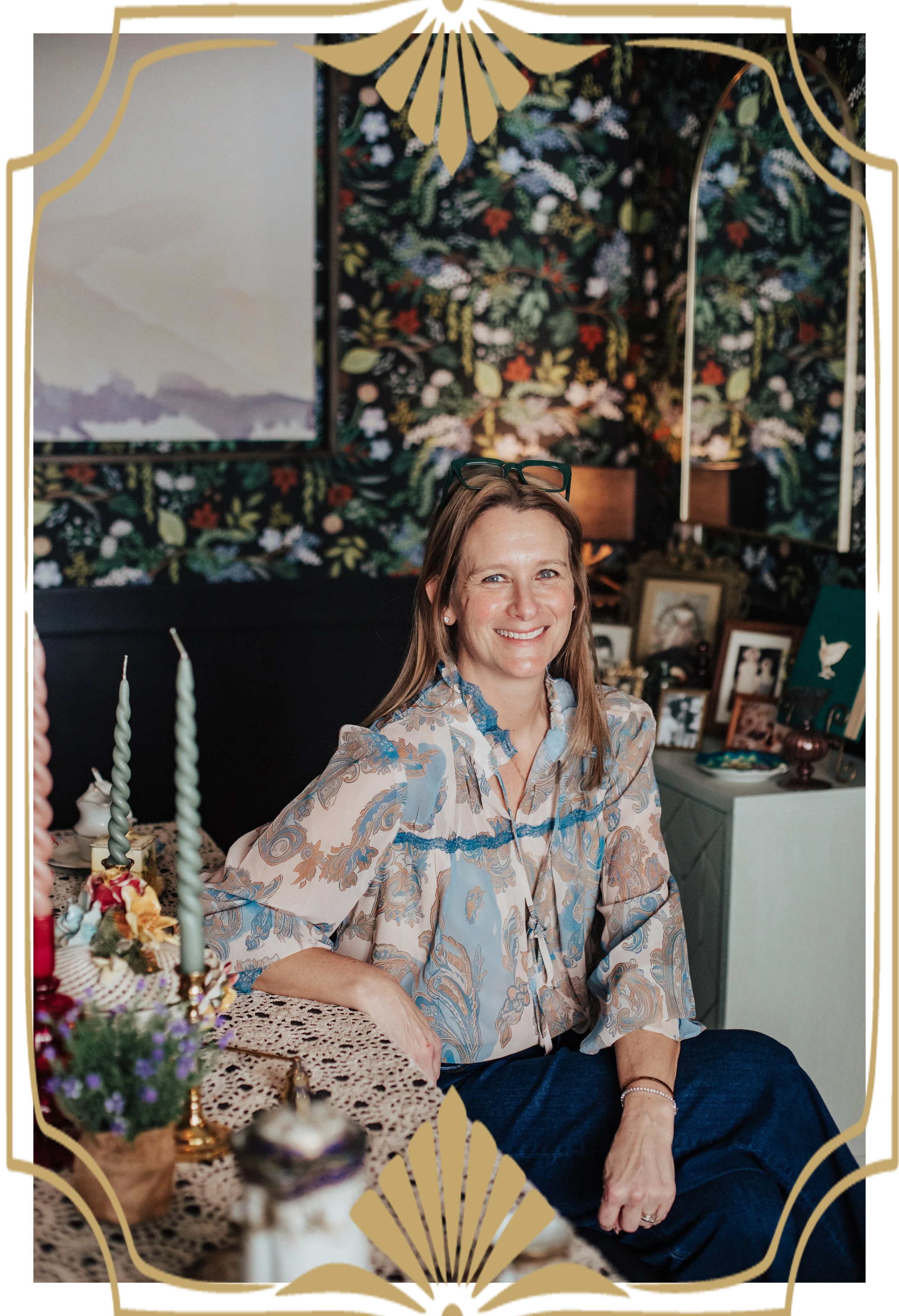 A woman with long brown hair and glasses on her head, smiling, sitting at a table with candles and floral decorations, in a room with floral wallpaper and framed photos in the background.