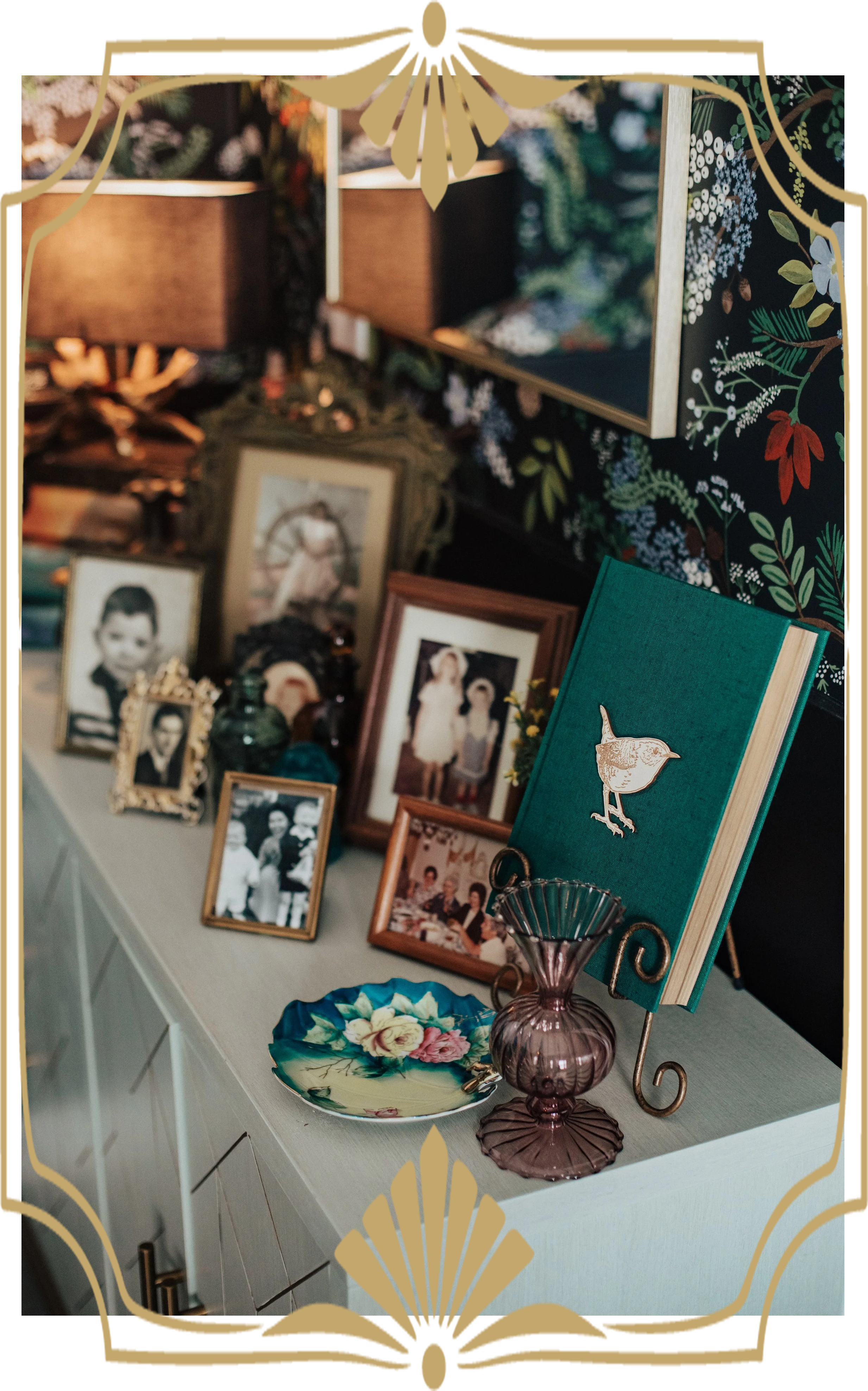 Display of framed black-and-white and color photographs, a decorative green book with a bird illustration, a pink glass vase, and a floral-patterned plate on a white sideboard, with a floral wallpaper in the background.