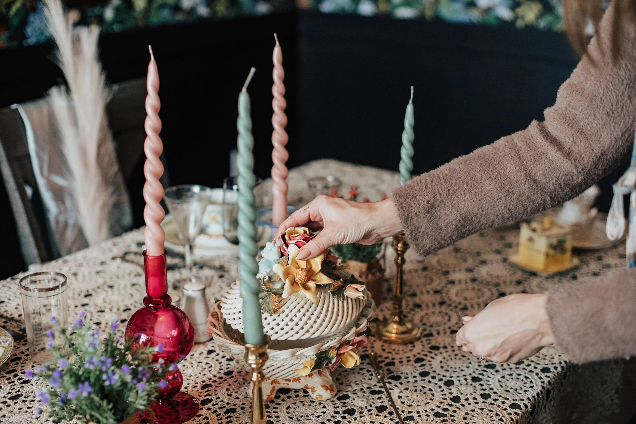 Person decorating a cake with flowers on a lace tablecloth, surrounded by candles and small plants.