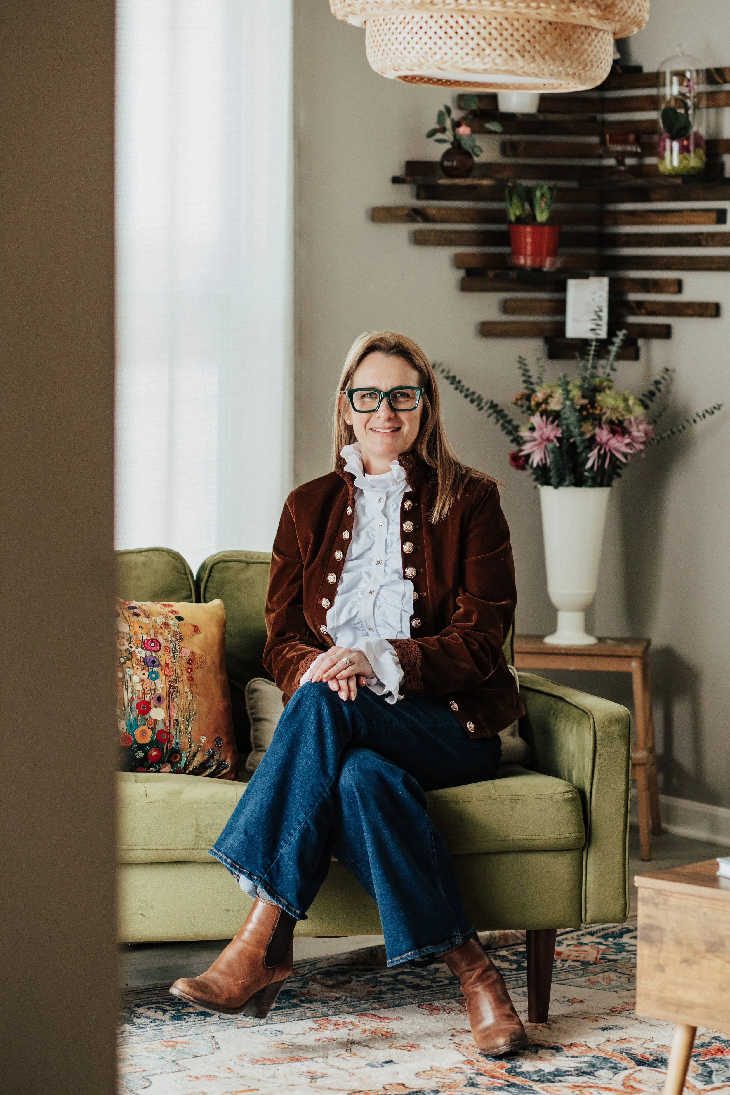 A woman with glasses and blonde hair sitting on a light green sofa in a cozy living room, smiling at the camera. She is wearing a white blouse, a brown jacket, and blue jeans. Behind her, there is a large white vase with pink and green flowers, a wooden wall decor piece with potted plants, and a curtain letting in natural light.