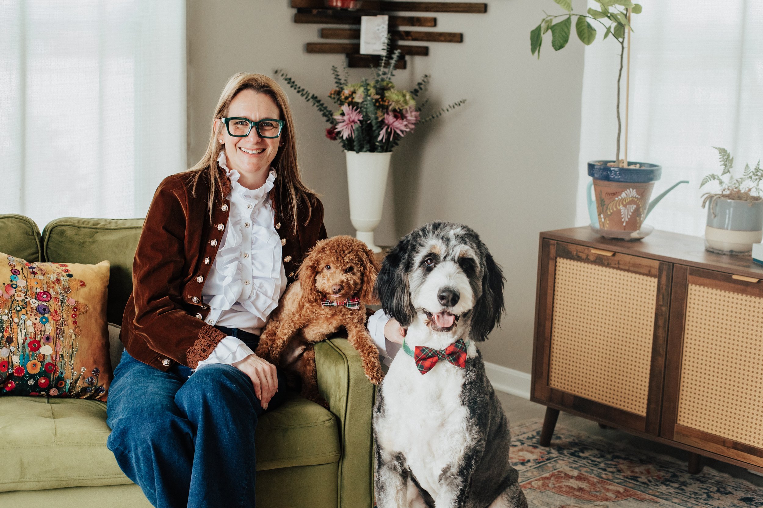 A woman with glasses sitting on a green couch, smiling, with two dogs: a small brown curly-haired dog on her lap and a large black and white dog with a red plaid bow tie sitting beside her in a cozy living room with plants and decorative pillows.
