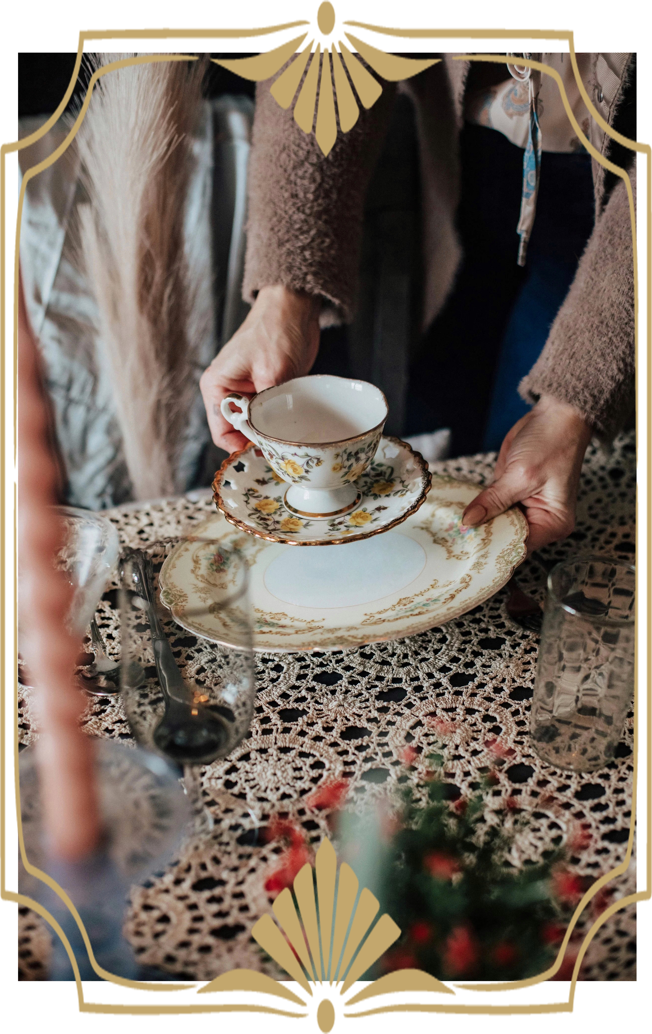 Close-up of a hand placing a floral-patterned teacup on a matching saucer on a lace tablecloth, with other glassware and blurred flowers in the foreground.