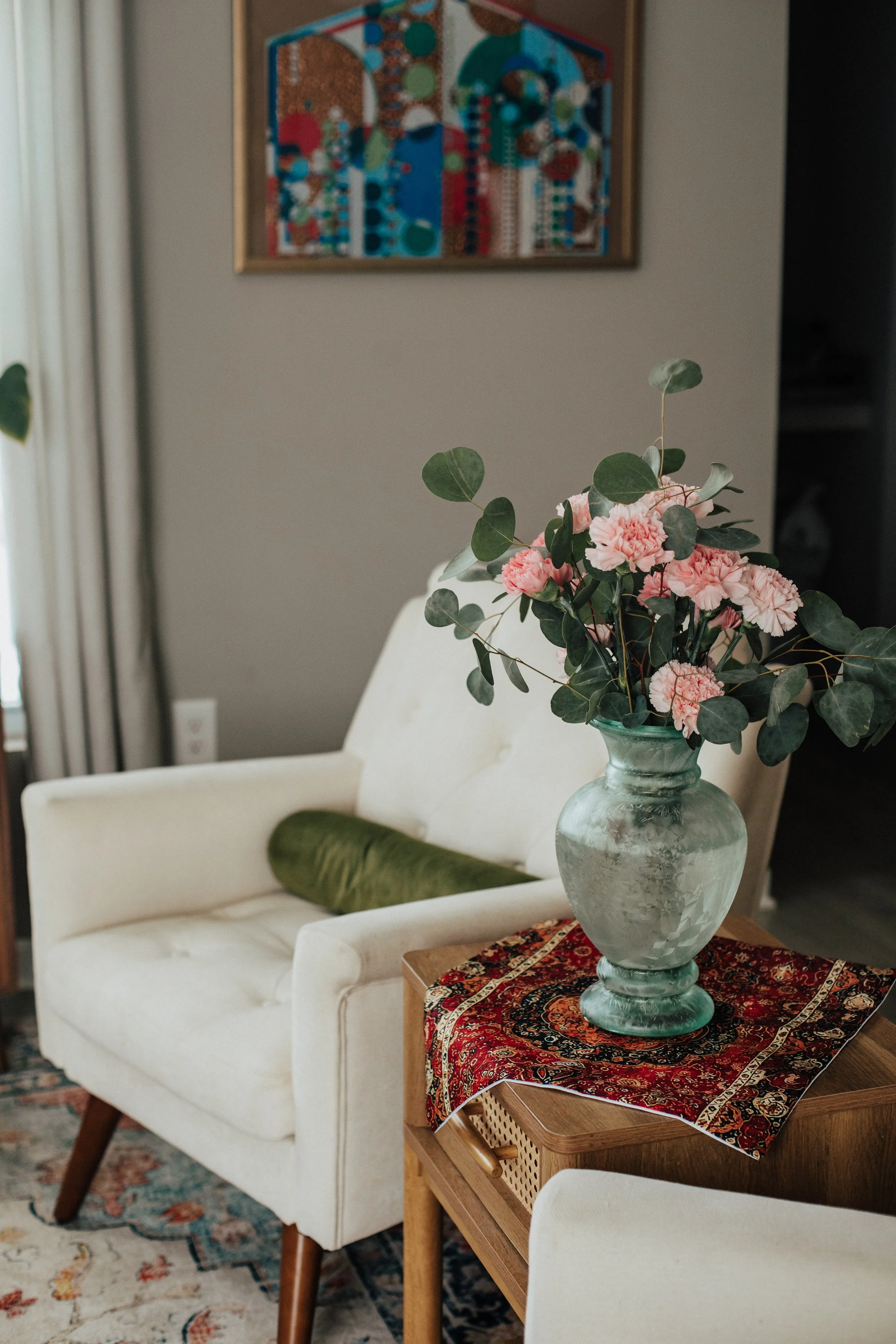 A living room with a white armchair, green pillow, wooden side table with a decorative fabric runner, and a large glass vase with pink carnations and eucalyptus, with a colorful abstract artwork on the wall in the background.