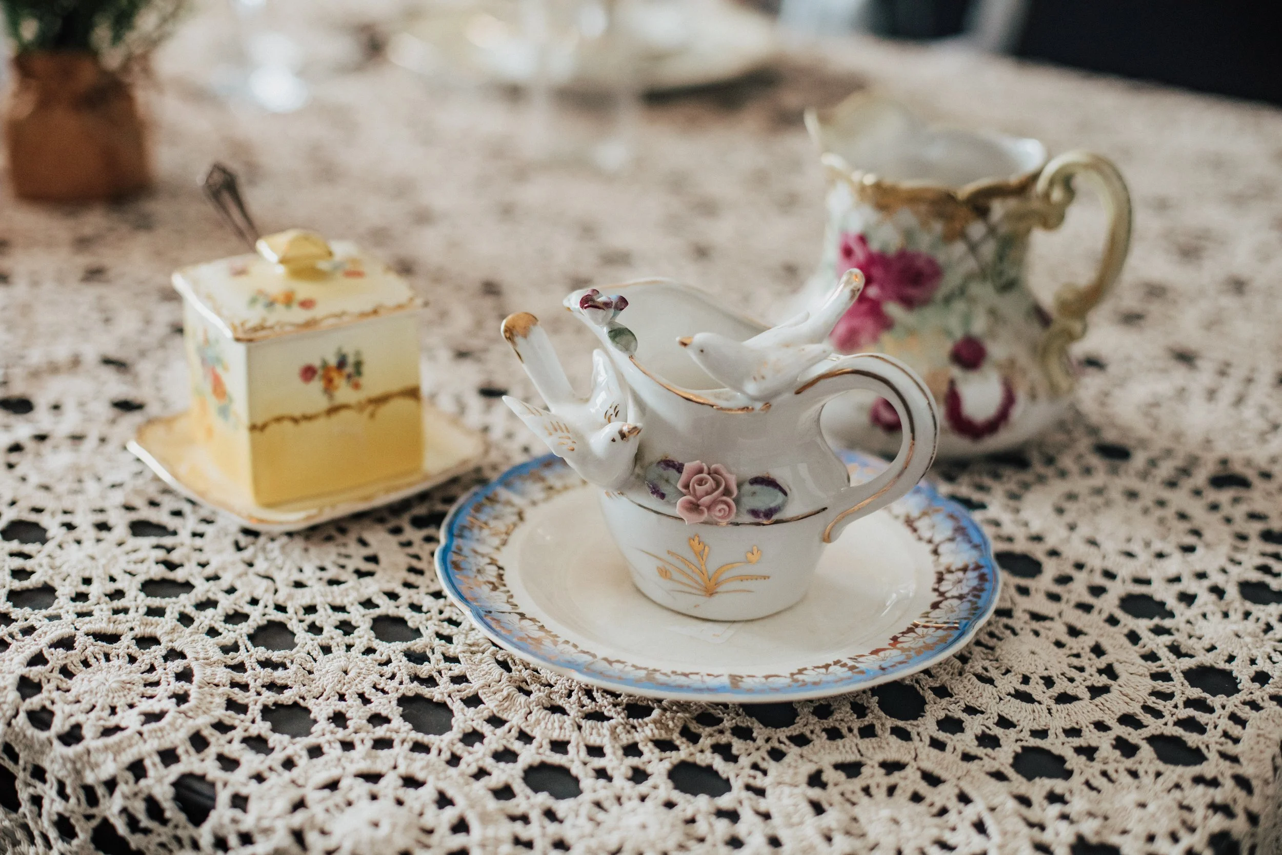 Vintage porcelain tea set with floral designs, including a teapot with bird figurines, a teacup on a matching saucer, and a small yellow box on a crocheted lace tablecloth.