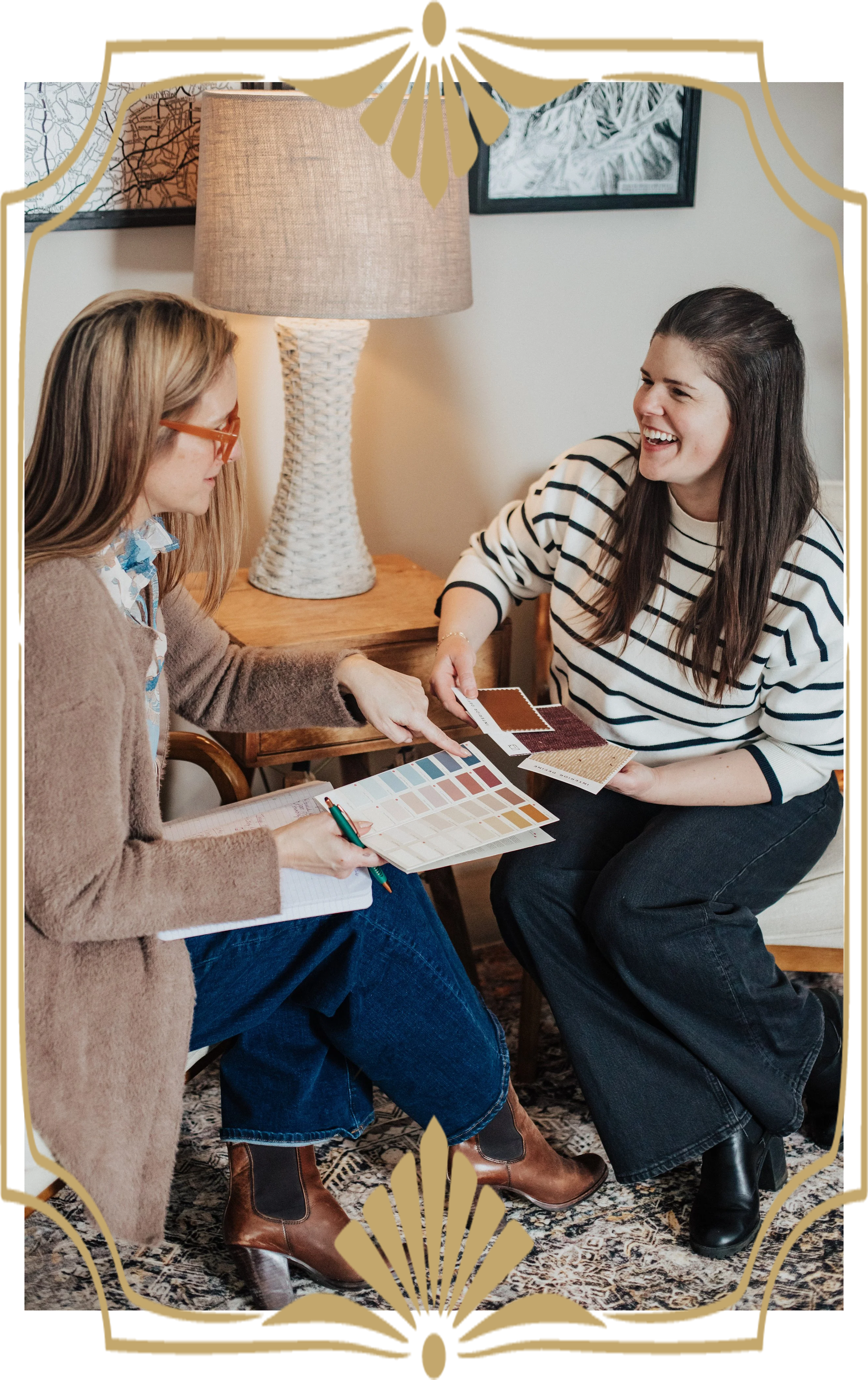 Two women sitting on a rug in a cozy room, smiling and sharing color swatches and design samples. One woman wears a striped shirt, the other a brown sweater. There is a lamp and framed artwork on the wall behind them.