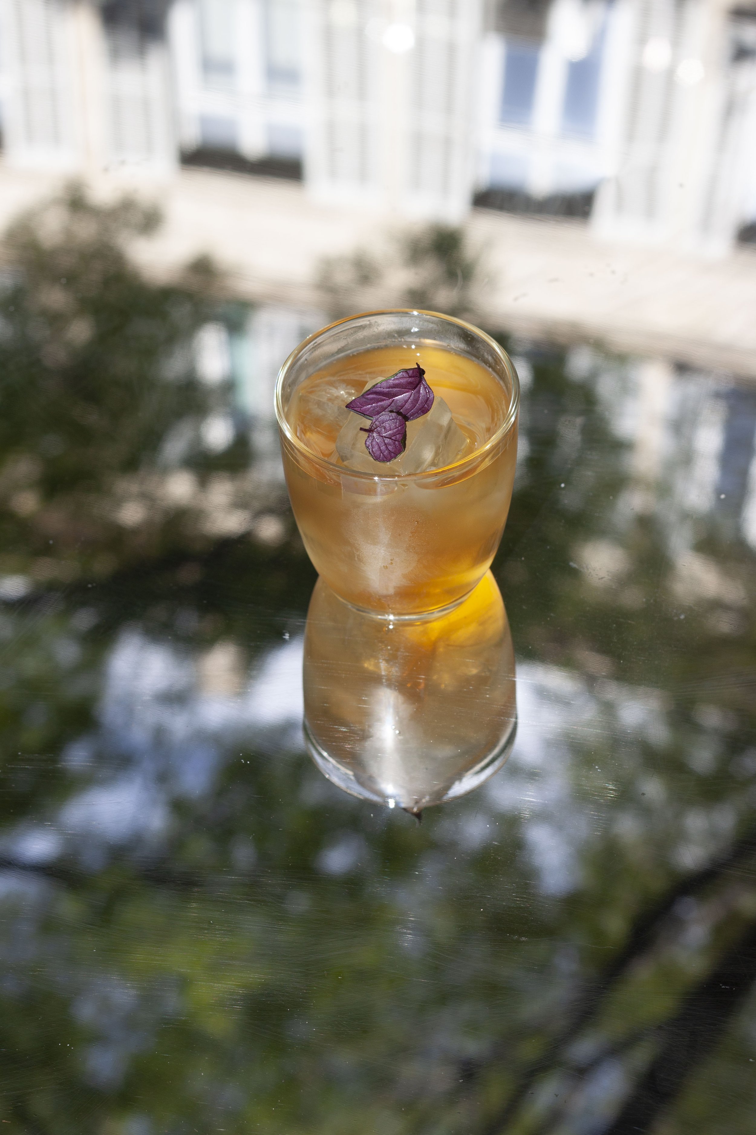 Un verre avec une boisson orangée, garnie de feuilles pourpres et de glaçons, placé sur une surface réfléchissante. En arrière-plan, fenêtres avec vue sur l'extérieur.