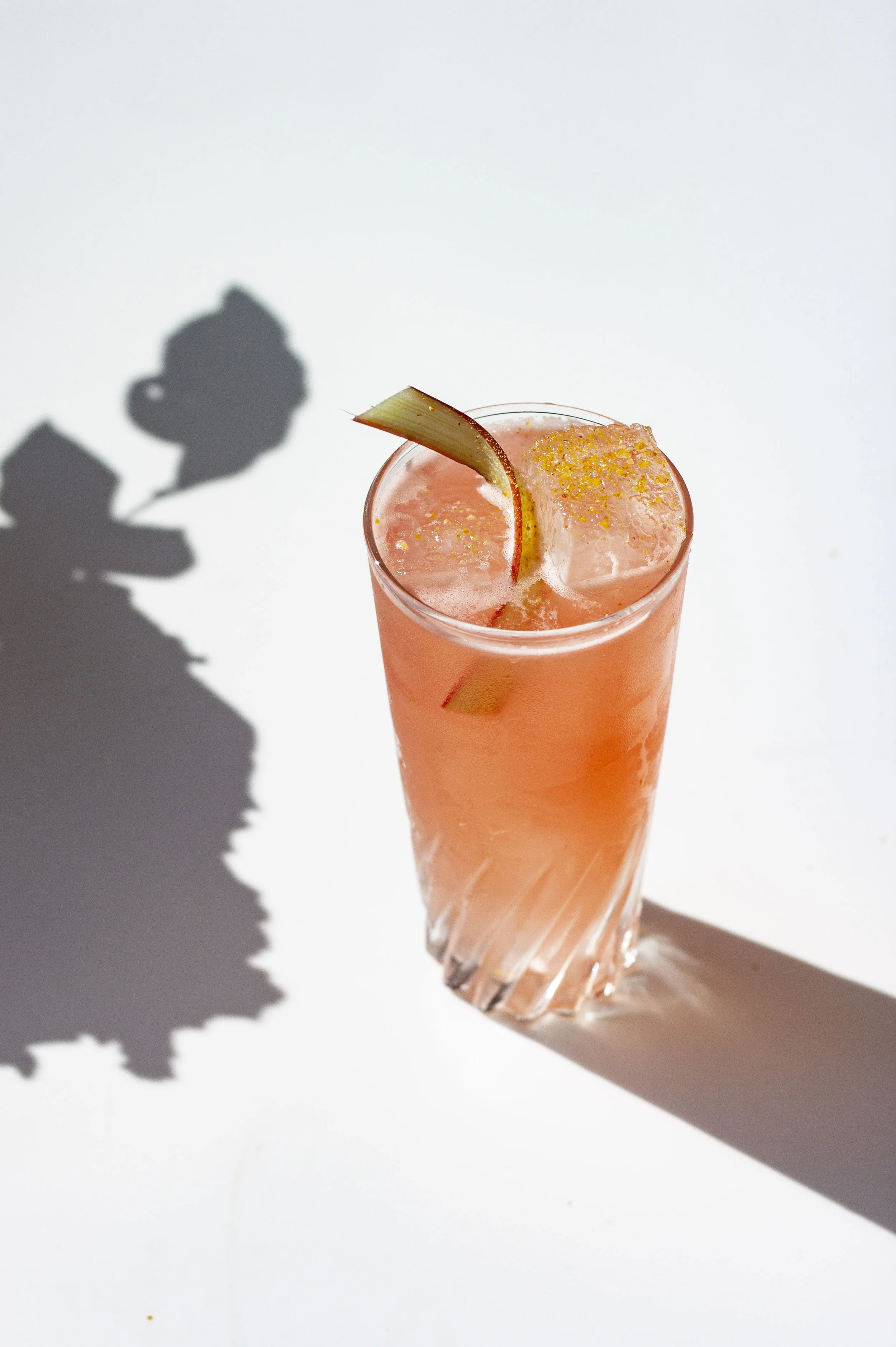 Un verre tallé contenant un cocktail rose avec des glaçons, garniture de tranche de fruit et paillettes d'or, avec une ombre de plante sur un fond blanc.
