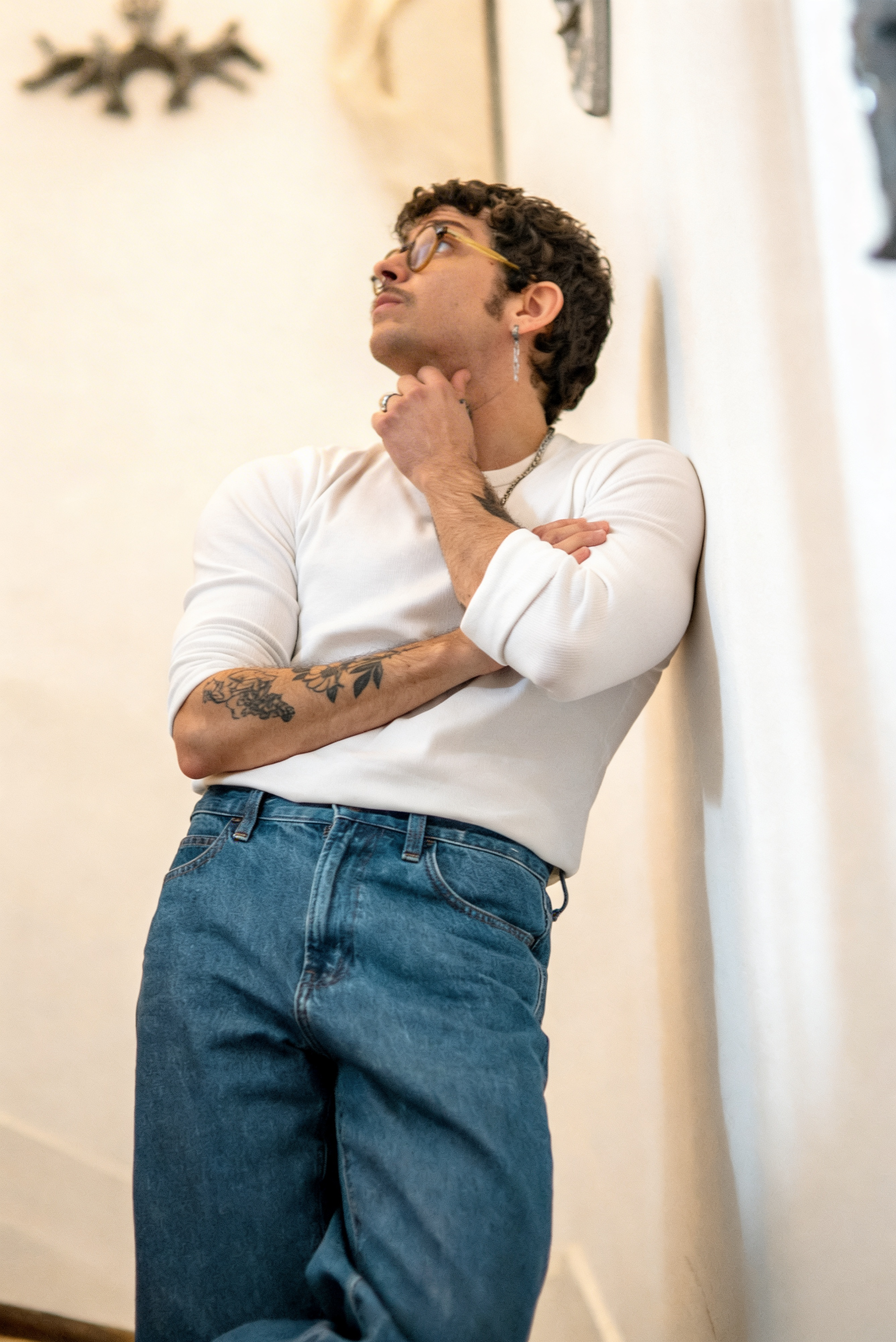 Jeune homme avec des cheveux bouclés, portant des lunettes, un earring, un t-shirt blanc et un jean, se tenant contre un mur, en train de réfléchir.