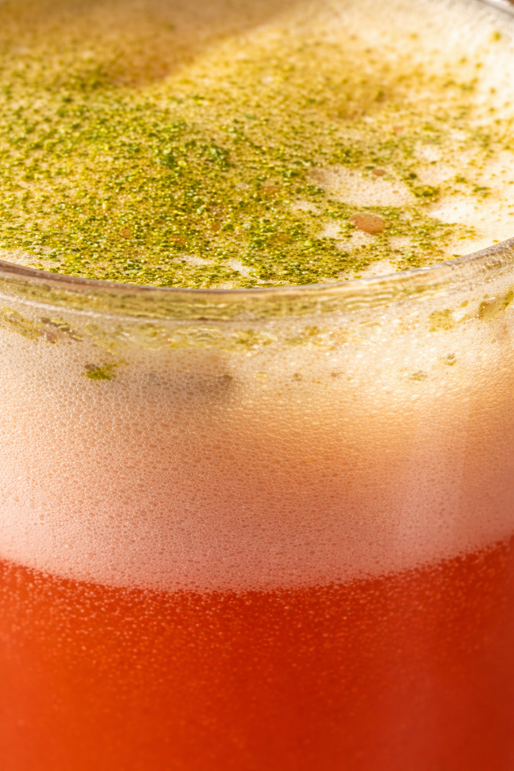 Une boisson dans un verre, avec une couche de mousse rose, un liquide rouge en dessous et une couche de mousse blanc avec du pollen ou des graines vertes sur le dessus.