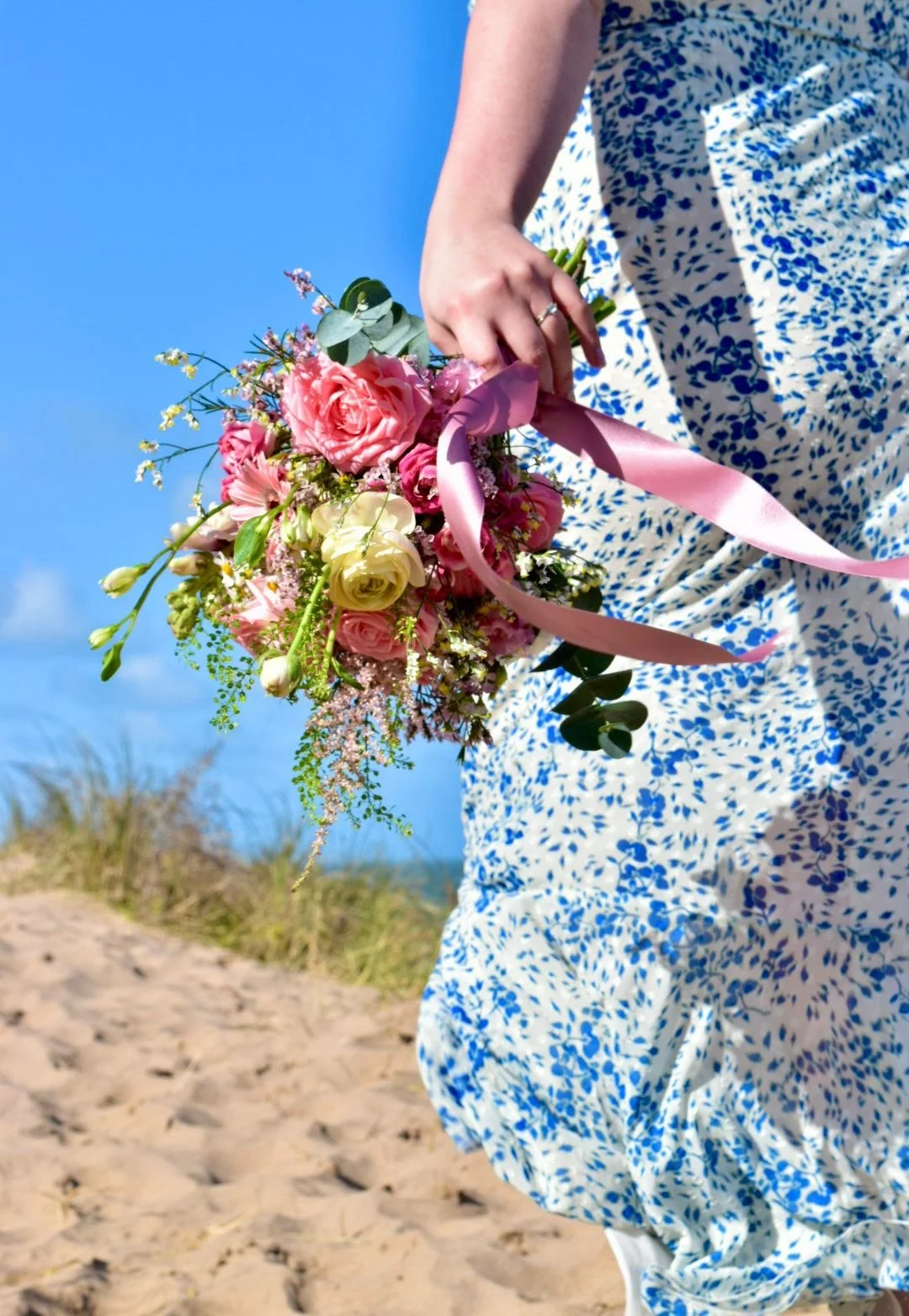 Person holding a bouquet of pink, white, and purple flowers, on a sandy beach under a blue sky.