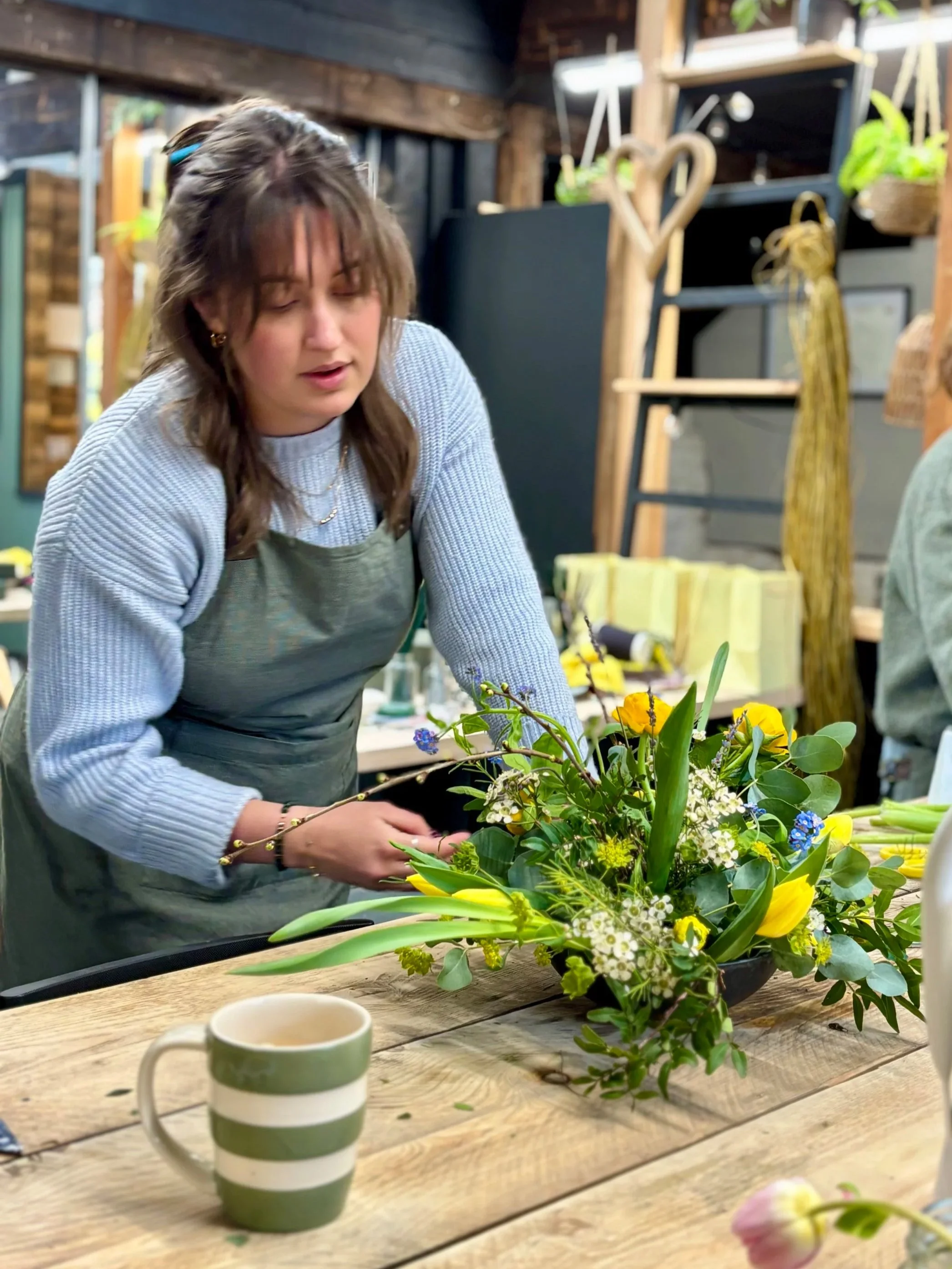 A woman arranging a floral centerpiece with yellow and white flowers in a black bowl, on a wooden table in Waterlooville.
