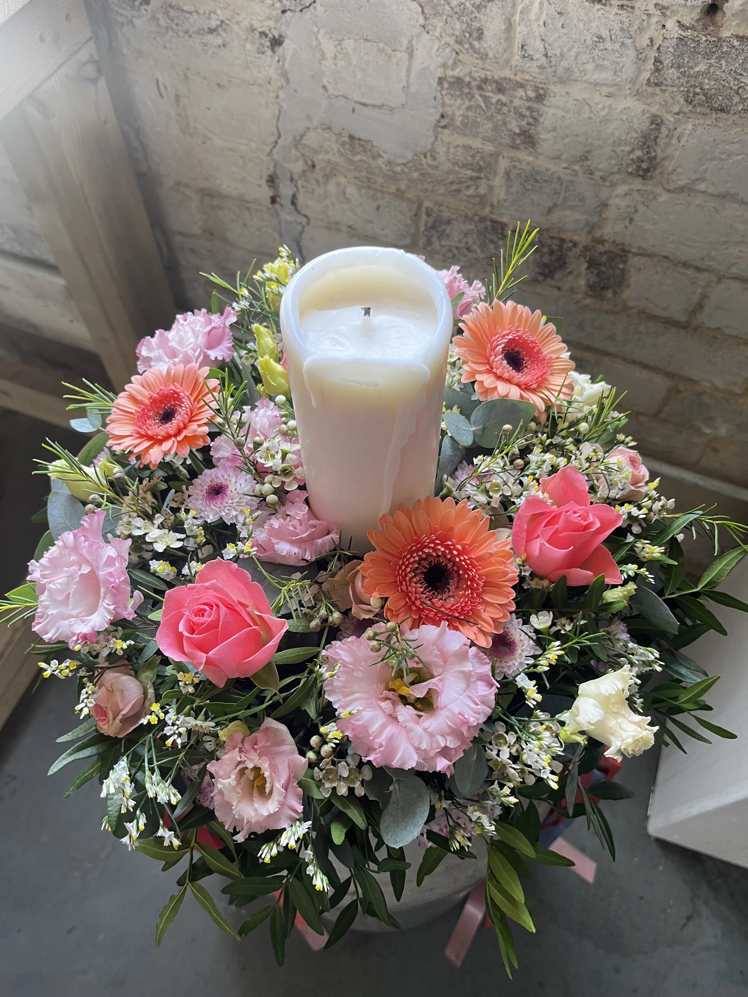 A floral arrangement with pink roses, orange gerbera daisies, pink carnations, white lisianthus, small white filler flowers, and green foliage surrounding a white candle, all placed on a gray surface against a brick wall background.