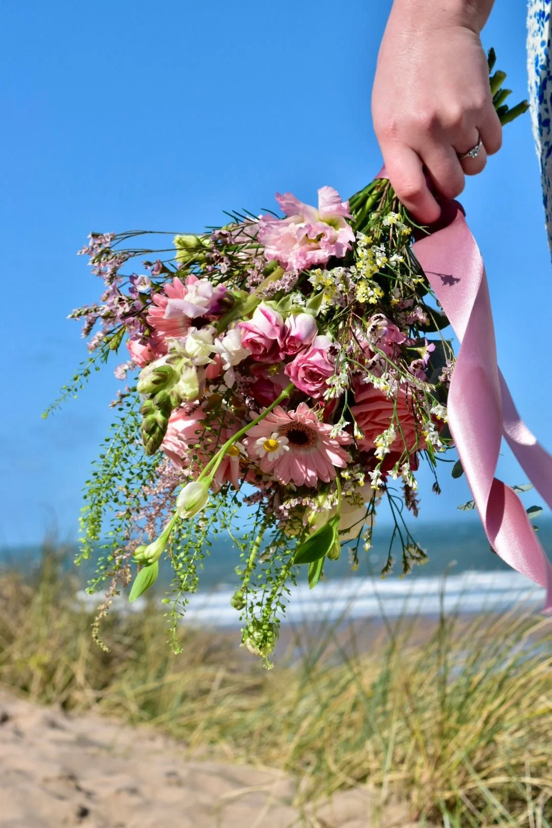 A person holding a bouquet of pink and white flowers with a pink ribbon, against a beach background with sand, grasses, and ocean under a blue sky.