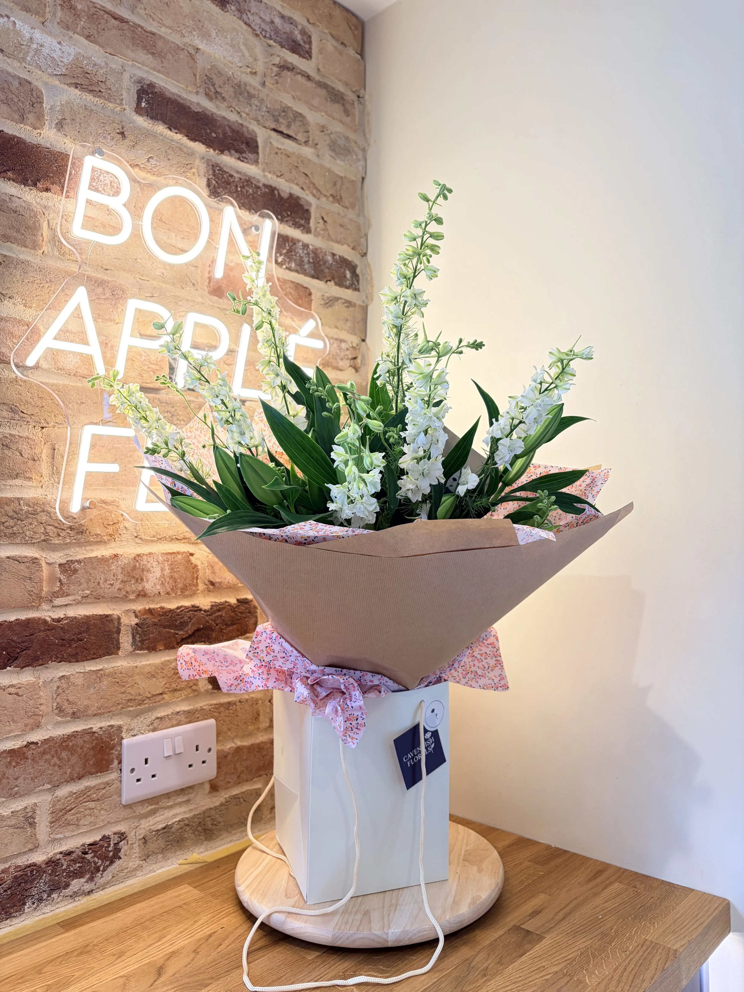 A flower bouquet with white flowers and green leaves on a white pedestal against a brick wall and white wall, with a neon sign reading 'BON APPETIT' in the background.