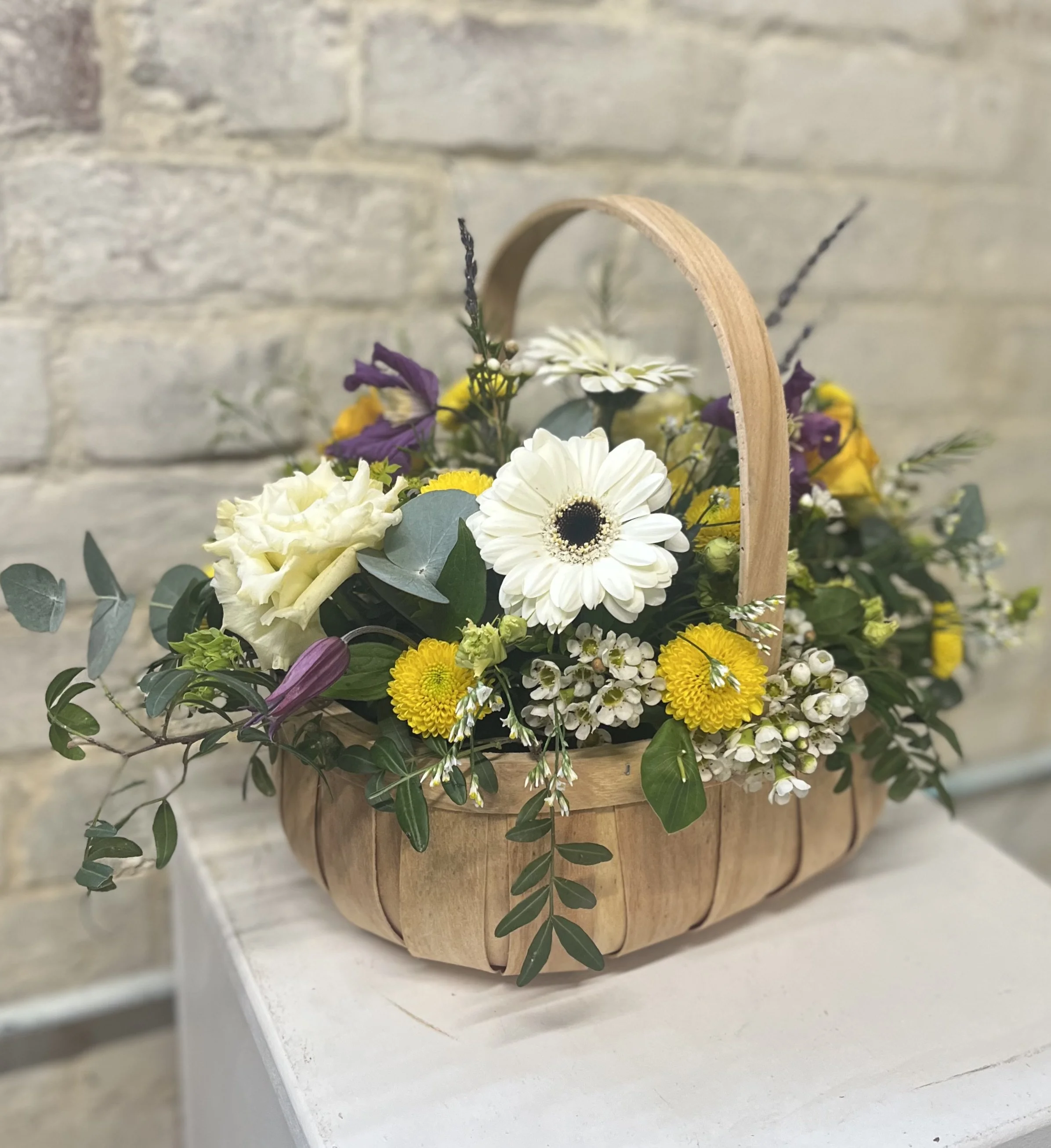 A wooden basket filled with white, yellow, purple, and green flowers and foliage on a light-colored surface against a brick wall background.