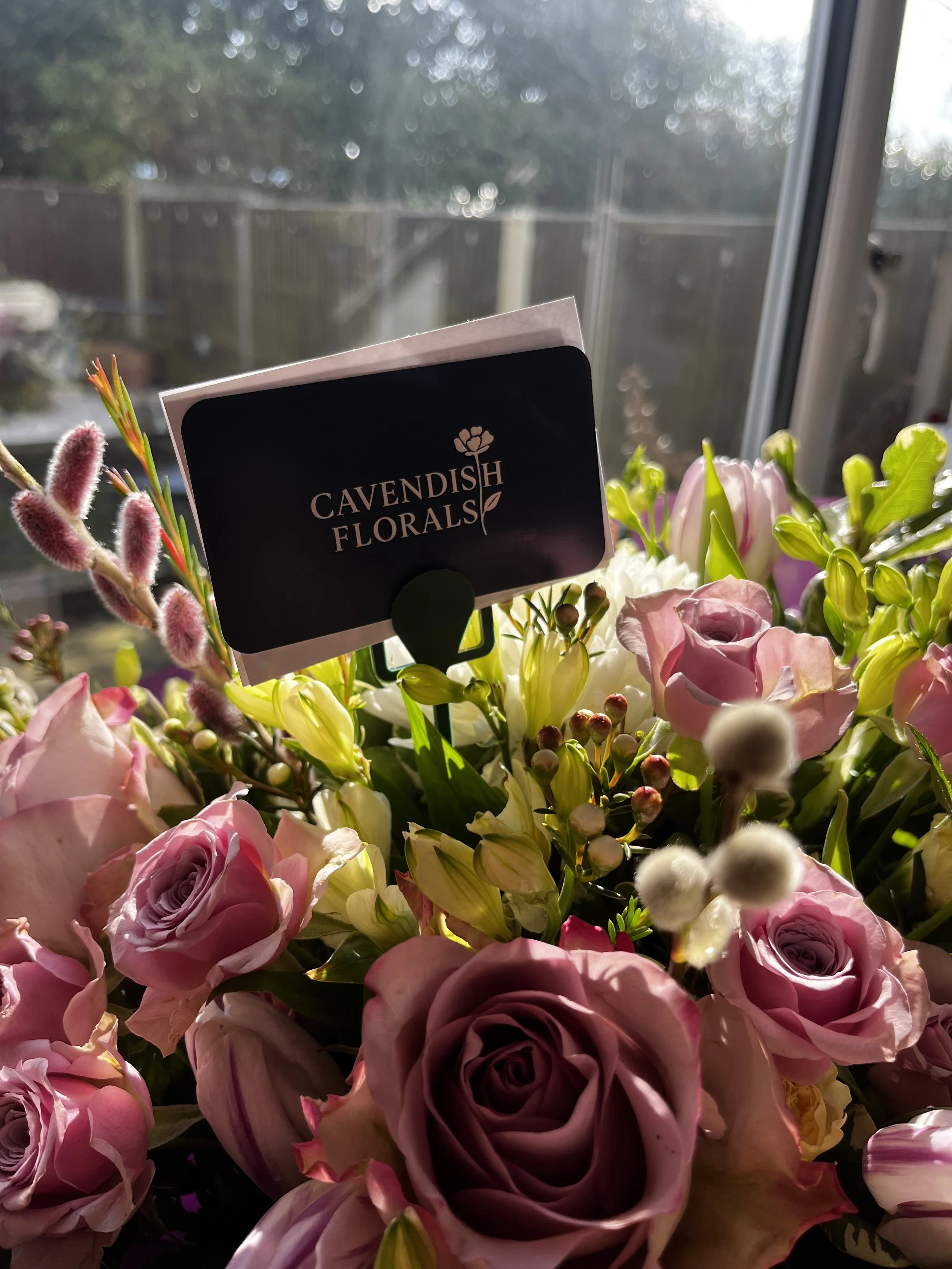Pink roses, white lilies, and other flowers in a bouquet with a black card that reads "Cavendish Florals" on a sunny windowsill.