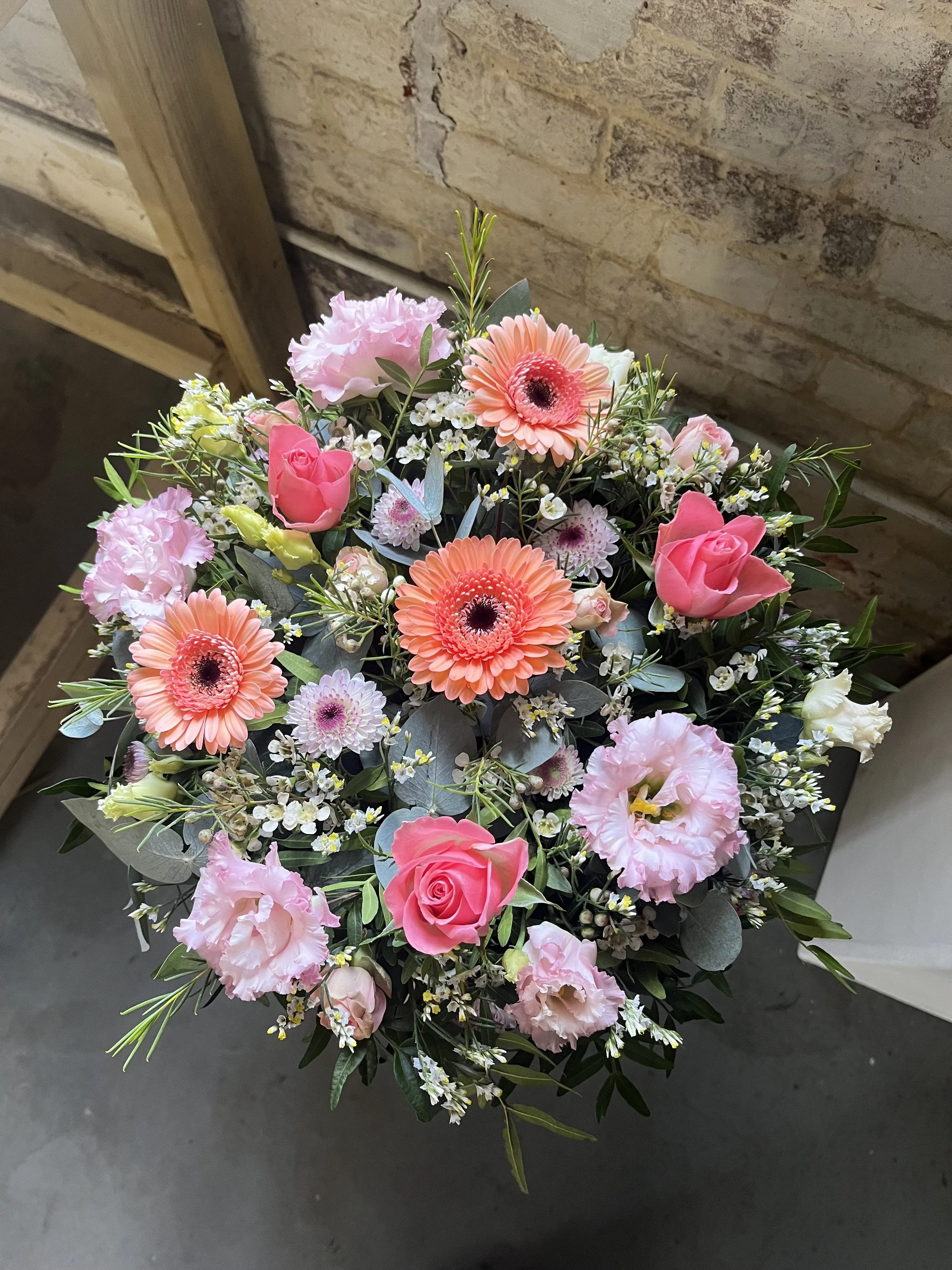 A bouquet of pink and peach flowers, including roses, gerbera daisies, carnations, and small filler flowers, arranged with green foliage.