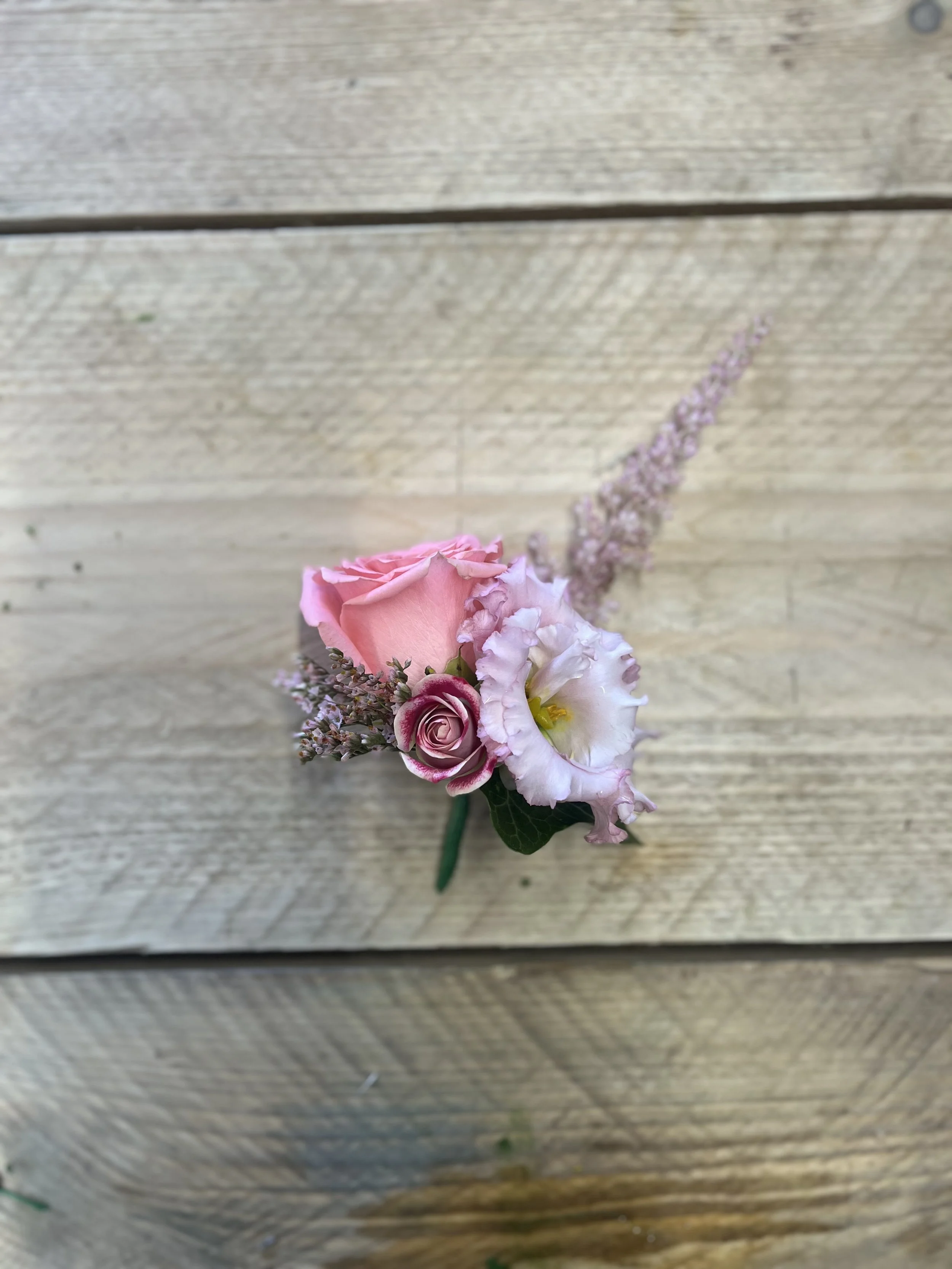 A boutonniere with pink roses, white lisianthus, and lavender accents on a wooden surface.