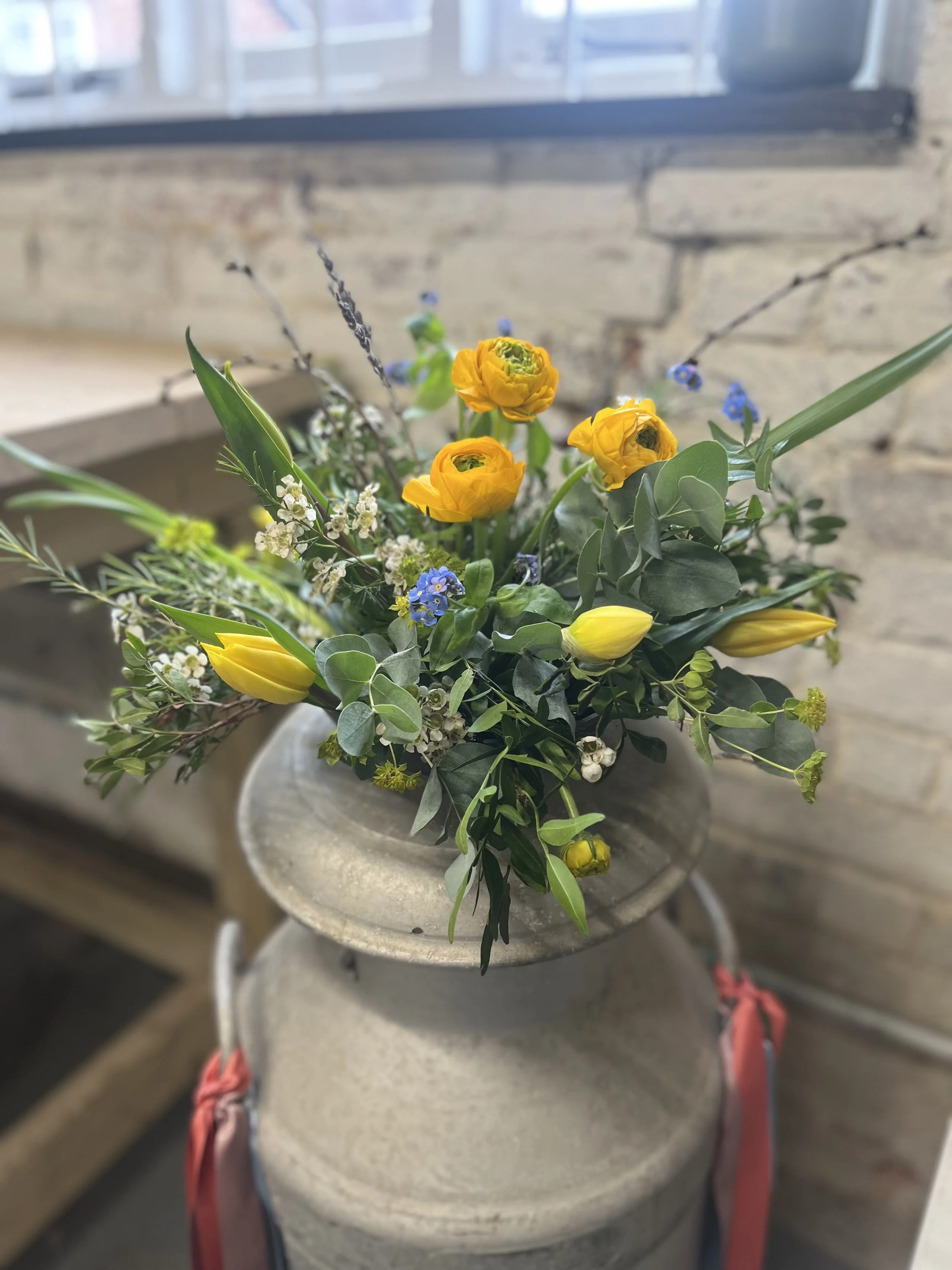 A bouquet of yellow flowers with green leaves and small blue and white accents in a large gray stone vase on a beige ledge against a brick wall and a window.