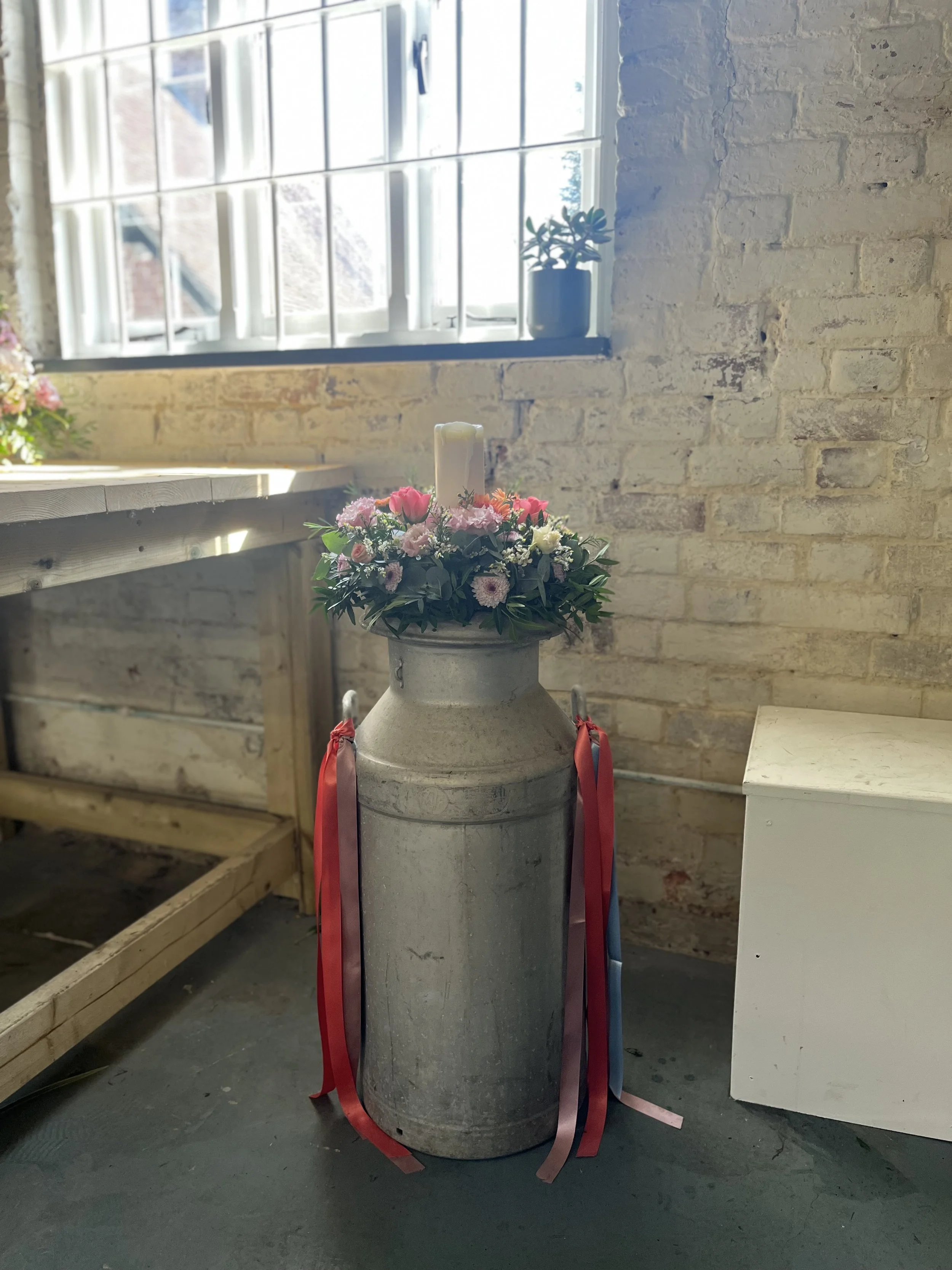 A large metal milk can with a floral arrangement including pink, white, and purple flowers, and a white candle on top, in an industrial-style room with a brick wall and a window.