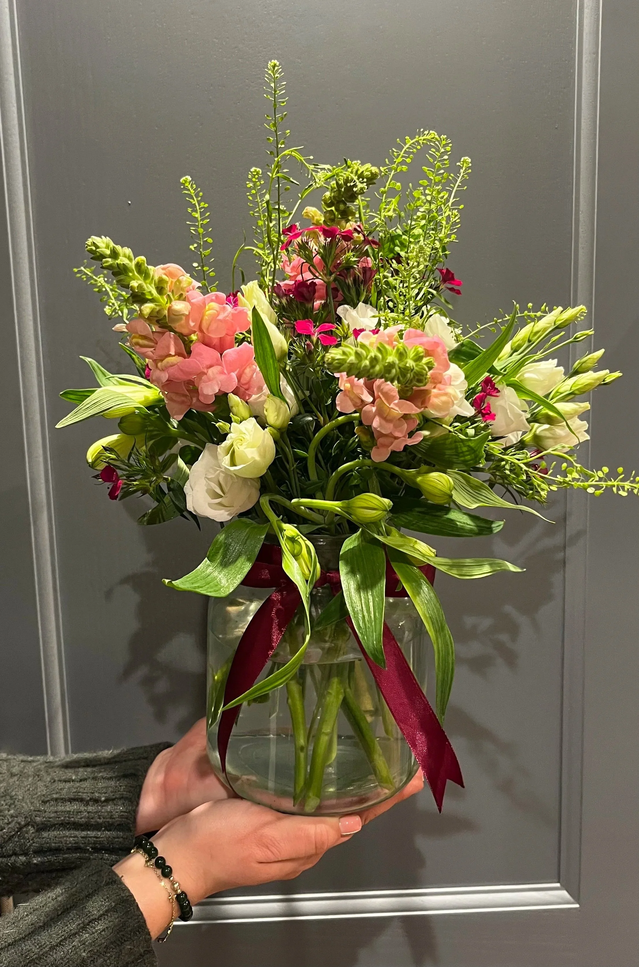 A person holding a glass vase with a colorful flower arrangement, including pink, white, and green flowers, tied with a dark red ribbon.