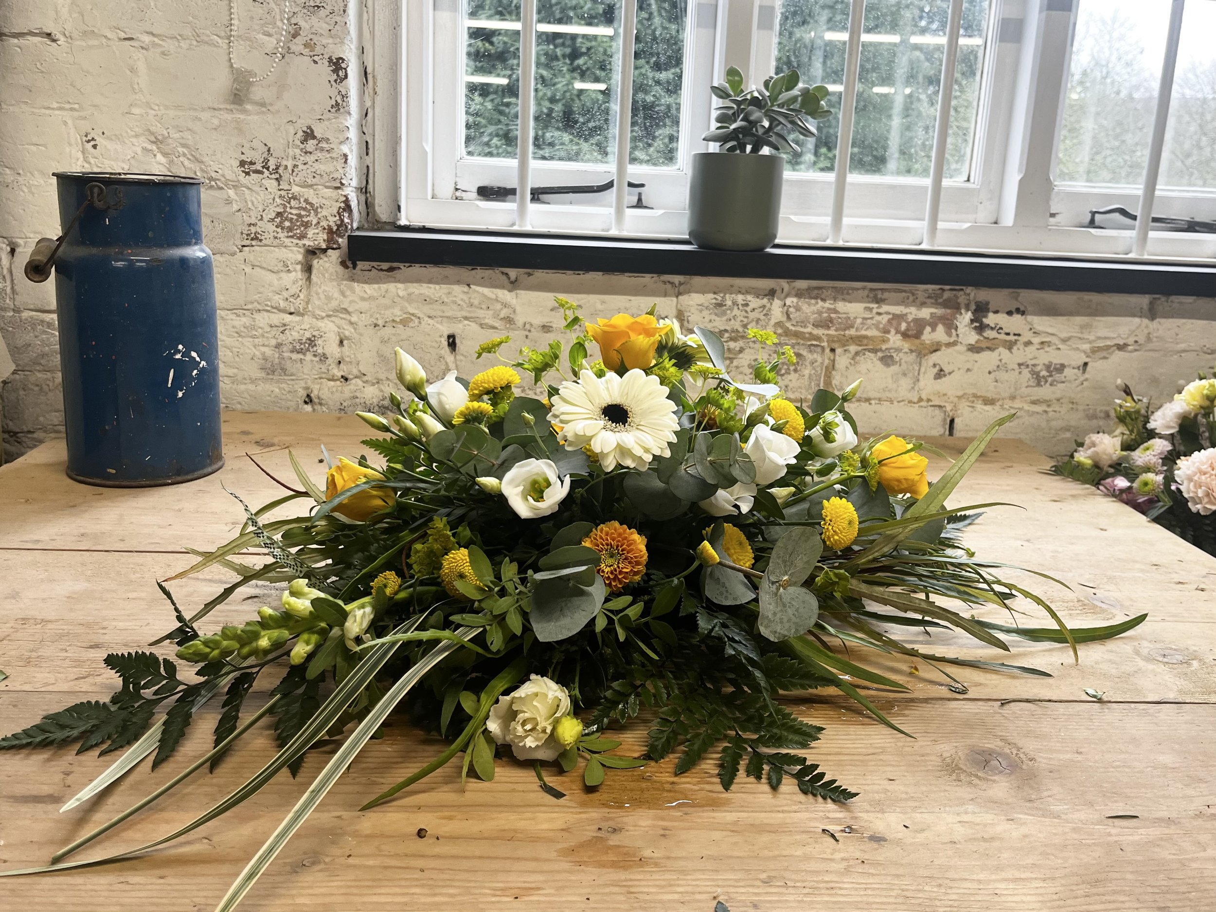 A floral arrangement of white, yellow, and orange flowers with green foliage, placed on a wooden table near a window with a brick wall background.