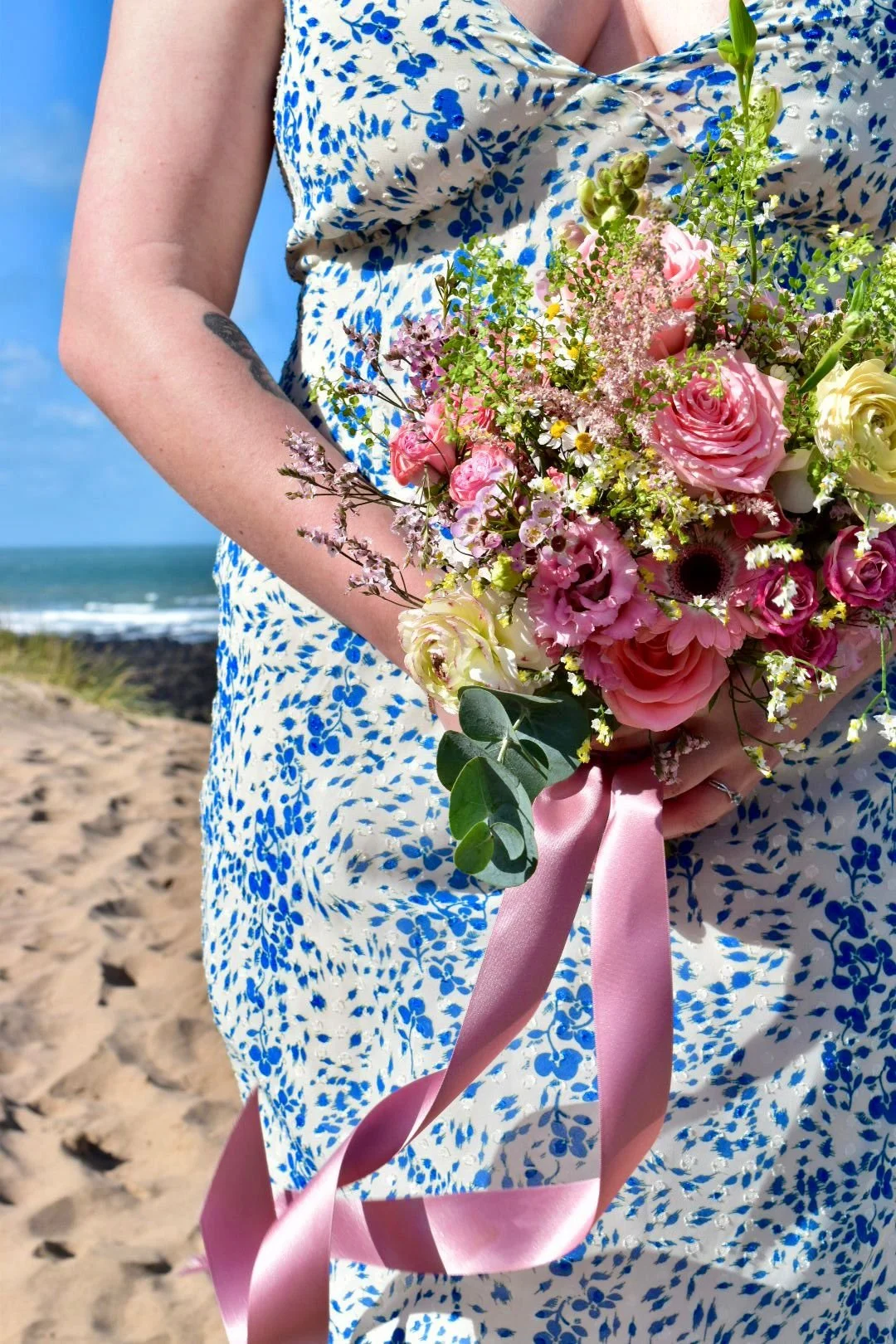 A person holding a bouquet of pink and white flowers with a pink ribbon, wearing a blue and white patterned dress, on a sandy beach with the ocean in the background.