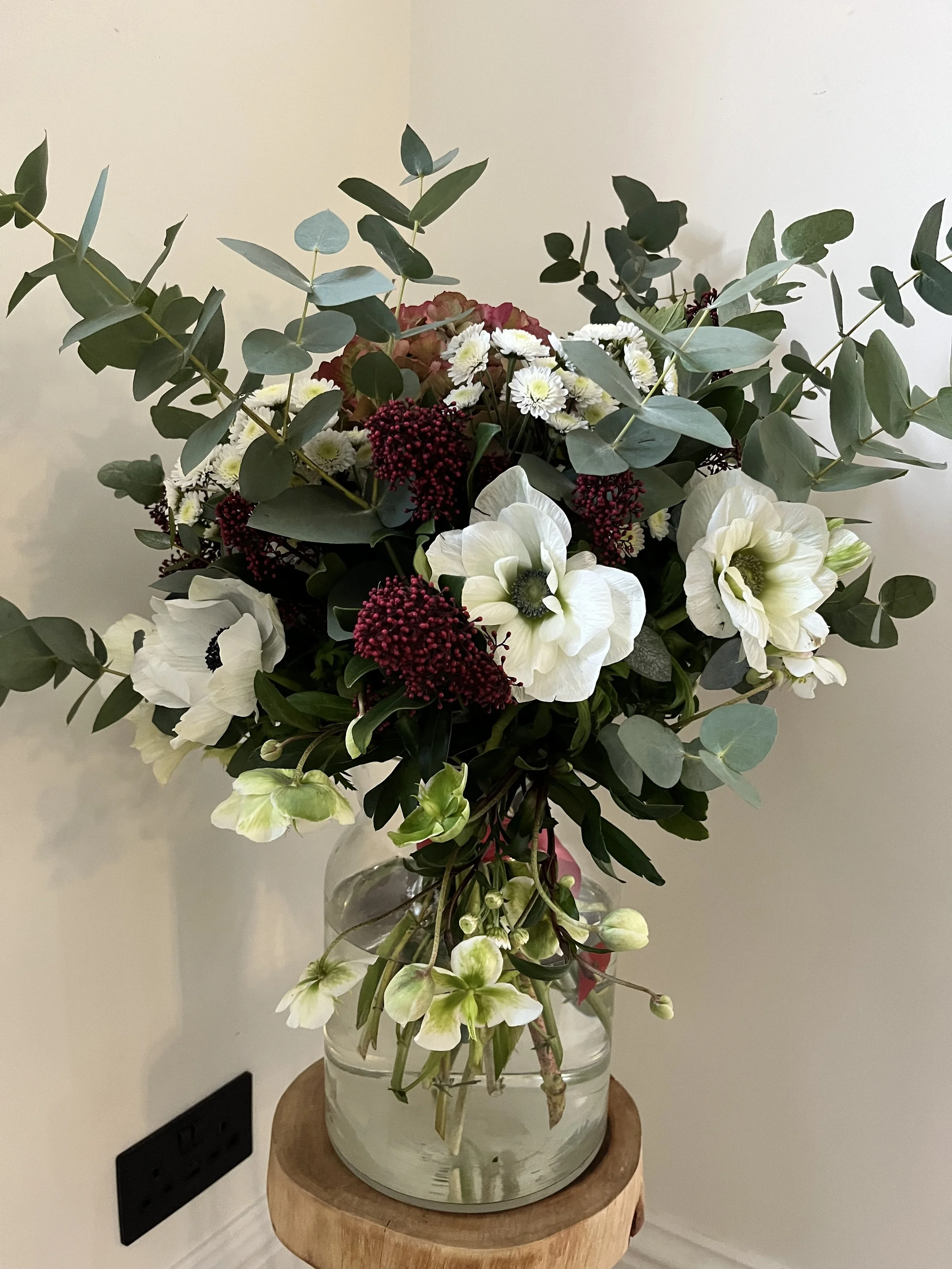 A bouquet of mixed flowers in a glass vase placed on a wooden stand against a plain wall, featuring white, cream, and burgundy flowers with green foliage.
